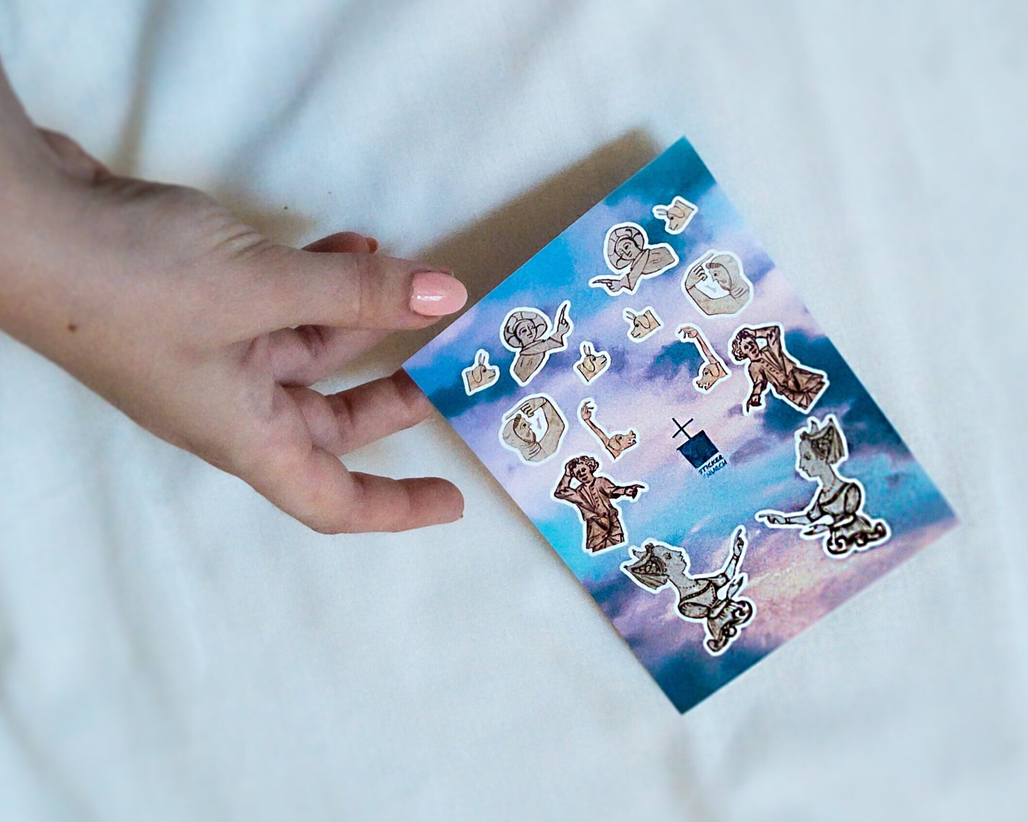 Hand with pale coral manicure holds a sticker sheet featuring designs of medieval manicules in the form of people and dogs, in front of a neutral background.