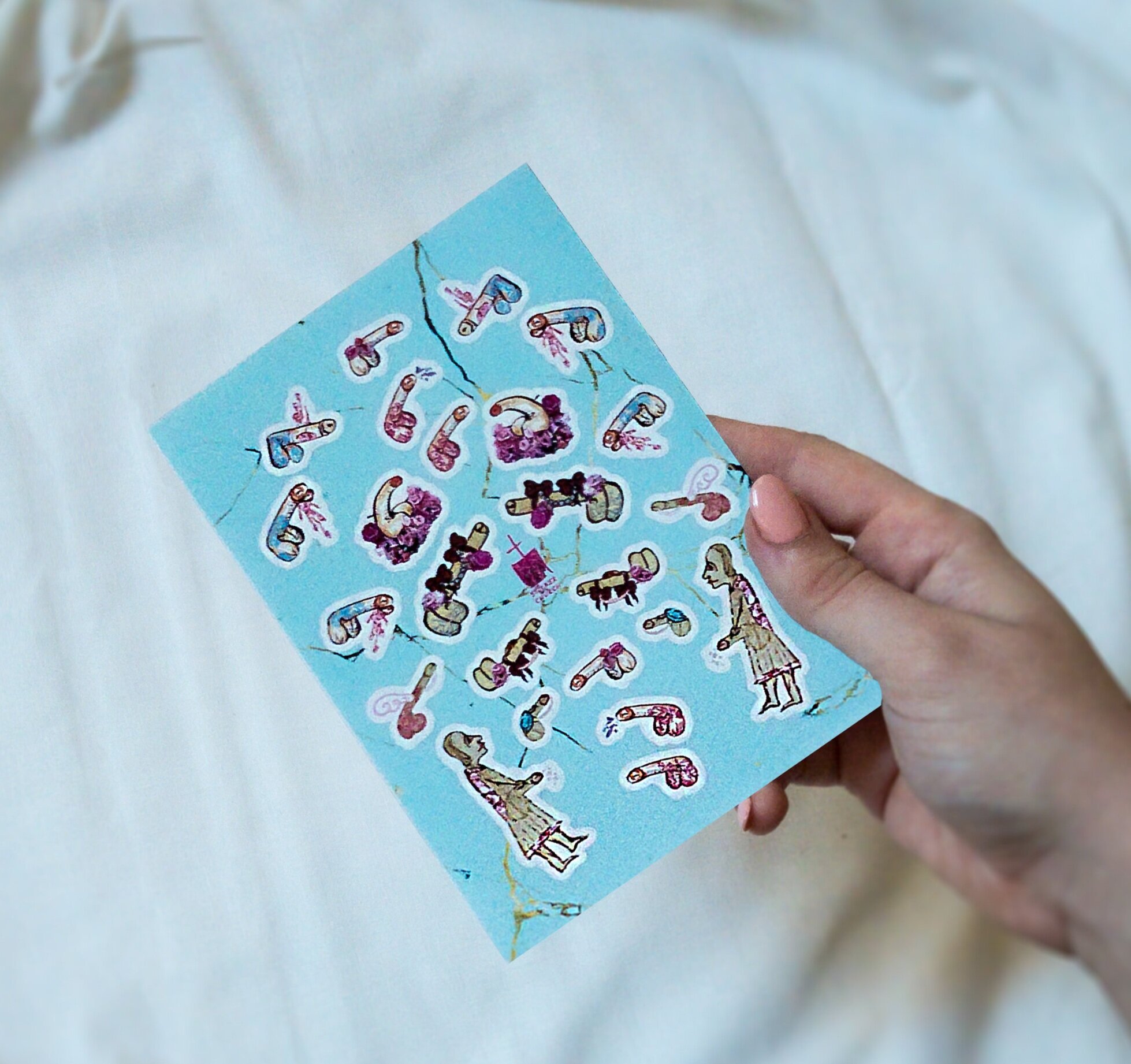 Hand with pale coral manicure holds a sticker sheet with designs of medieval penicule (penis-manicule) stickers, in front of a neutral background.