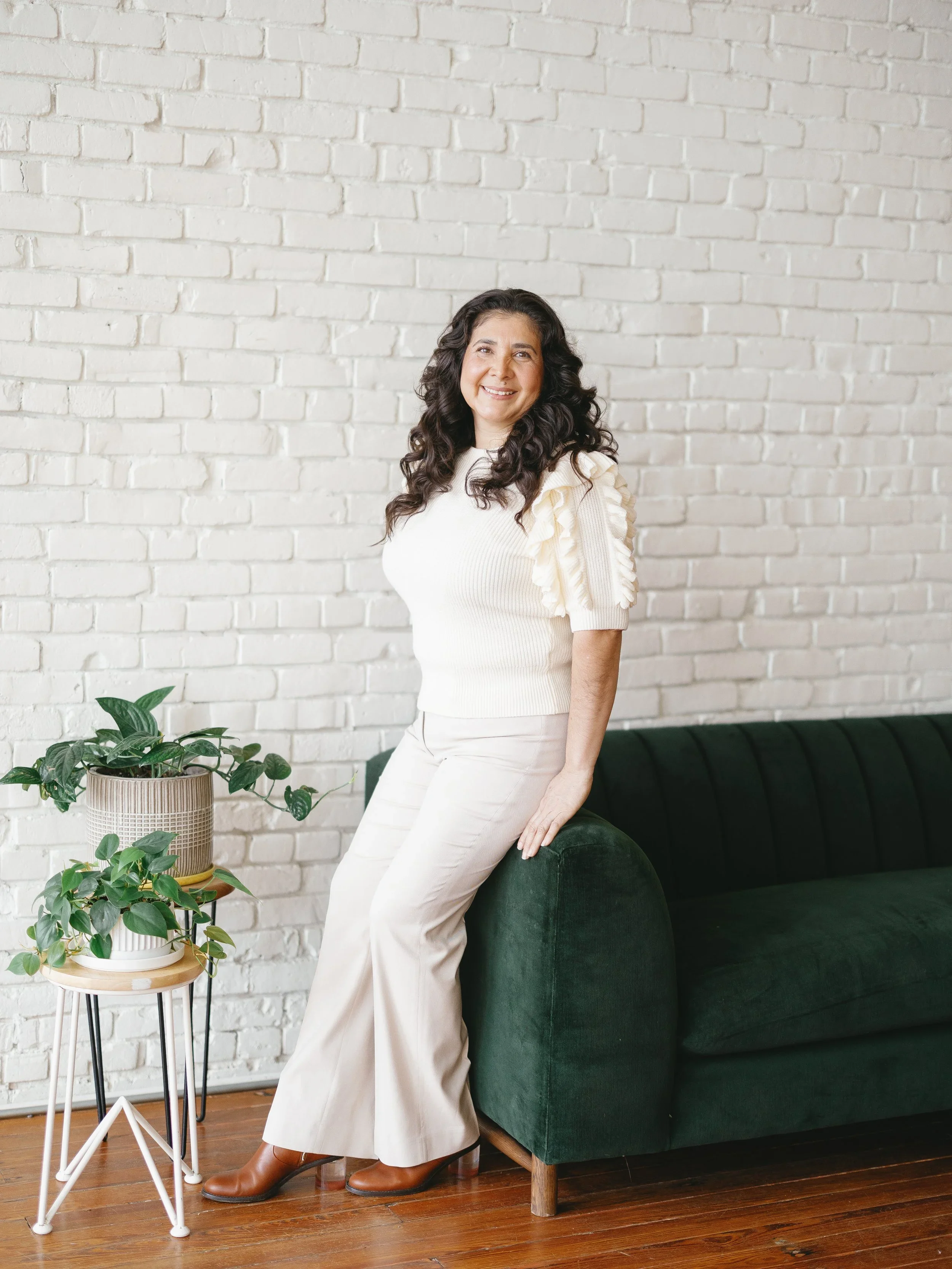 A woman with long, wavy brown hair sitting on a white stool in front of a white brick wall, smiling at the camera near a large window.
