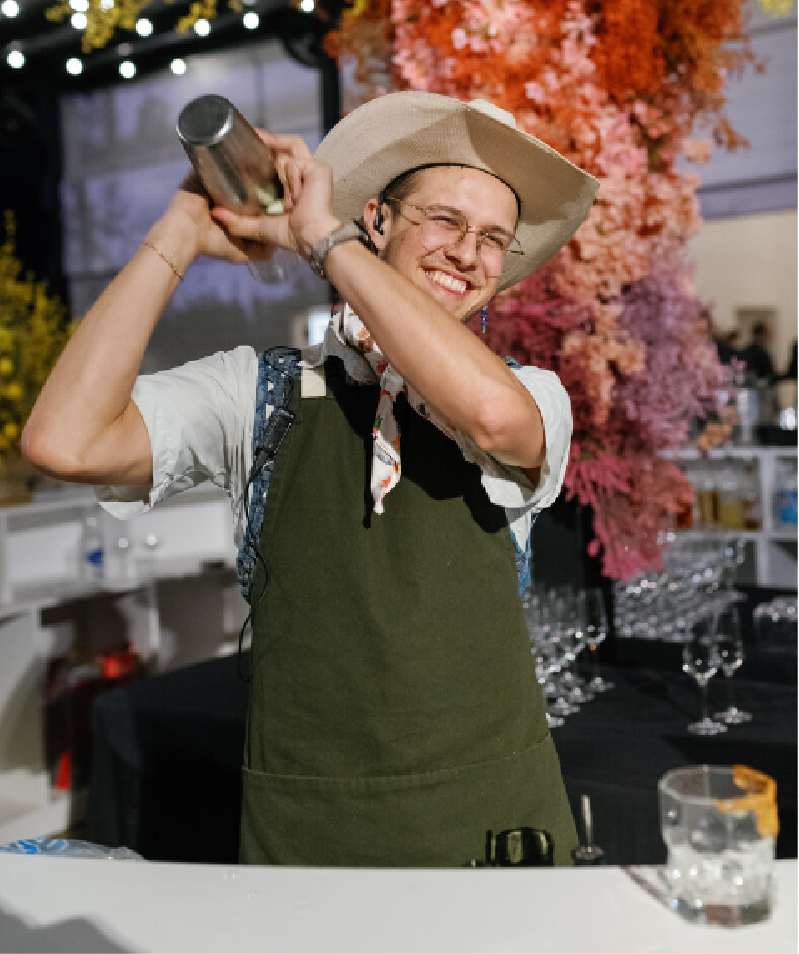 A smiling person in a white shirt and green apron with a large straw hat, holding a cocktail shaker above their shoulder in a festive setting with colorful flowers in the background.