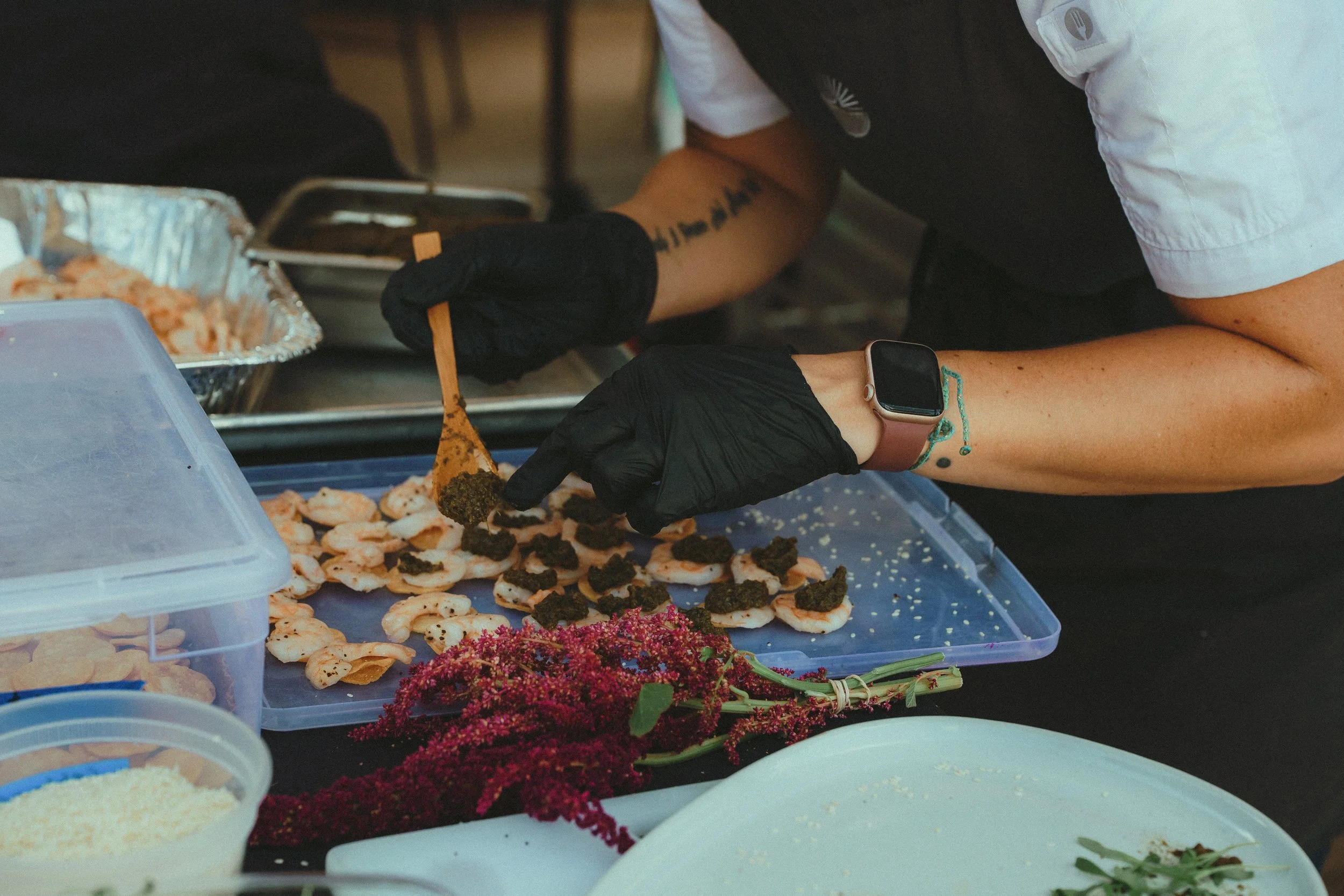 A person wearing black gloves and a smartwatch is preparing shrimp with a spoon on a busy kitchen or food stall. There are trays of shrimp, green herbs, and other ingredients on the table.