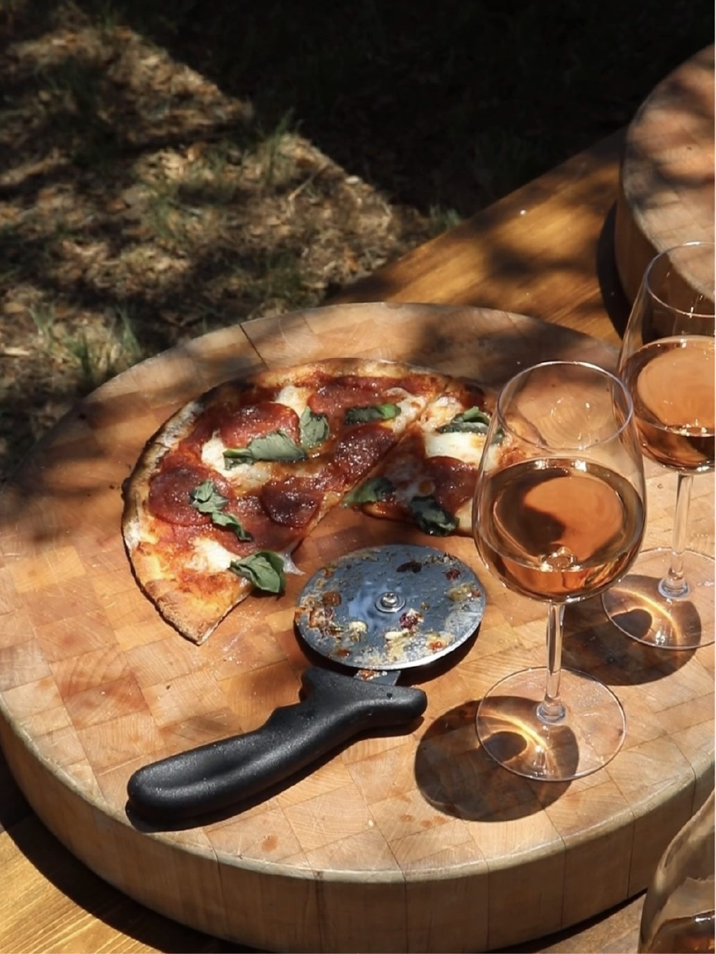Half-eaten pizza with basil leaves on a wooden pizza peel, with two glasses of rosé wine on a wooden table outside.