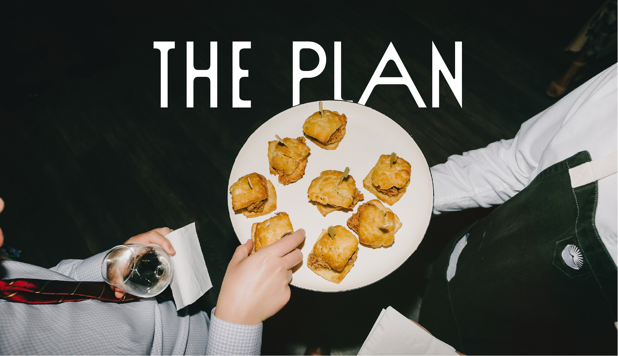 Overhead view of a waiter holding a white plate with ten mini fried chicken and biscuit sliders garnished with small skewers. A person in a gray shirt is holding a glass of white wine. The words "THE PLAN" are overlaid at the top of the image.