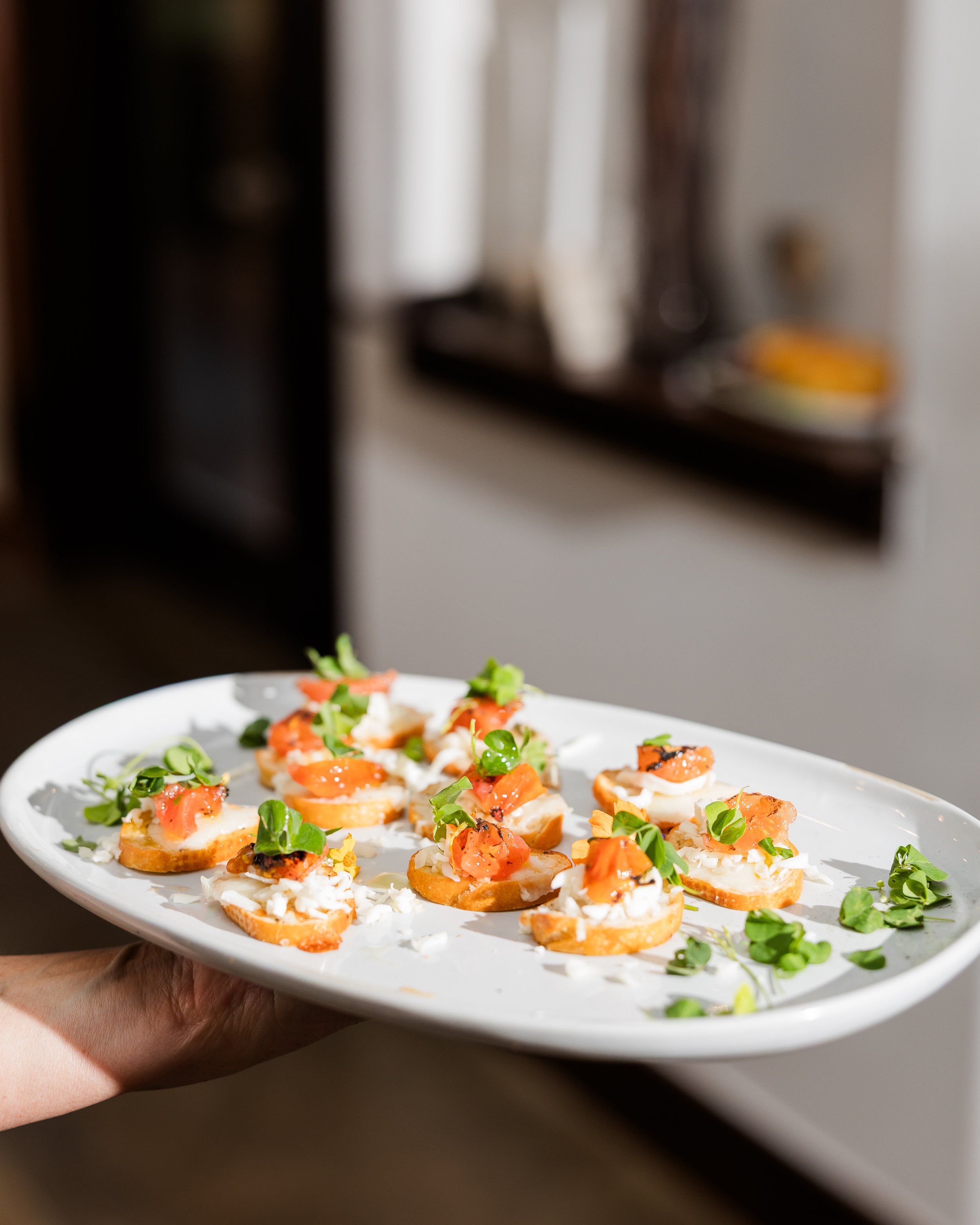 A hand holding a large white plate with small appetizers topped with smoked salmon, cream cheese, and garnished with microgreens, set in a bright room.