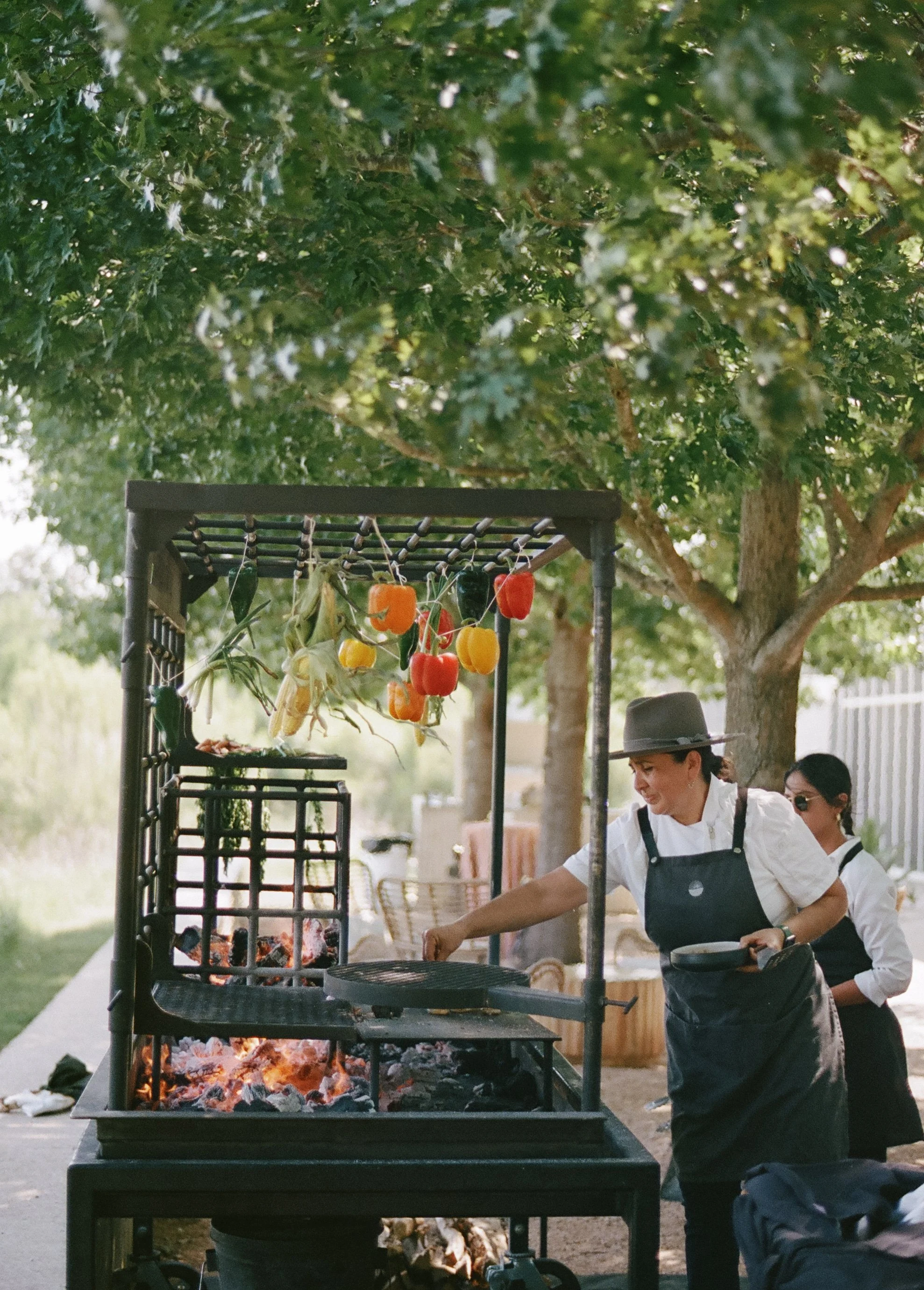 Two women cook outdoors over a grill with hanging vegetables. They are under trees, with one woman reaching towards the grill and the other standing nearby.