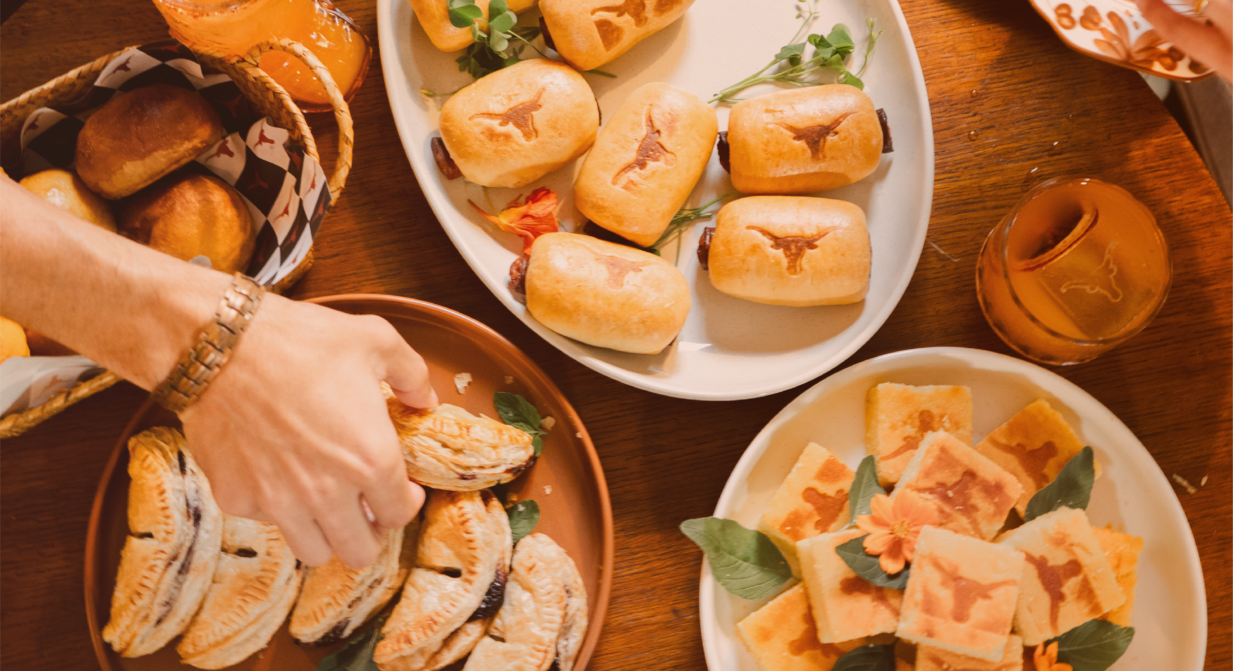 A table with various foods including sandwiches with the Texas Longhorn logo, slices of cake with a flower decoration, and a basket of baked goods.