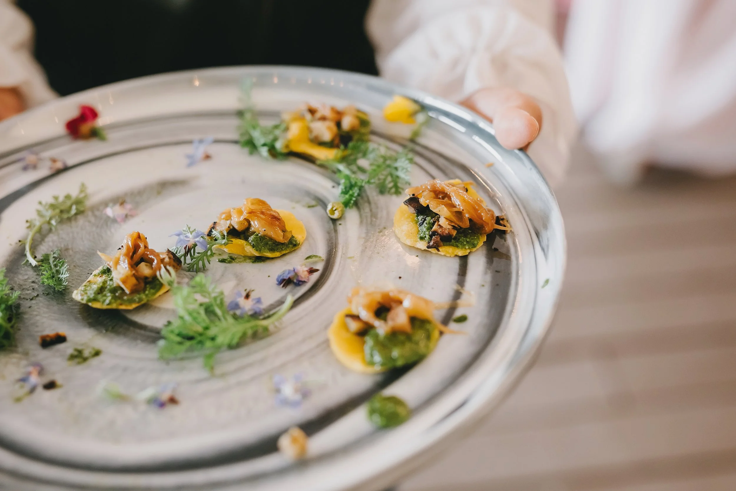 Close-up of a silver platter with small, colorful food garnished with fresh herbs and edible flowers, held by a person in a white shirt.