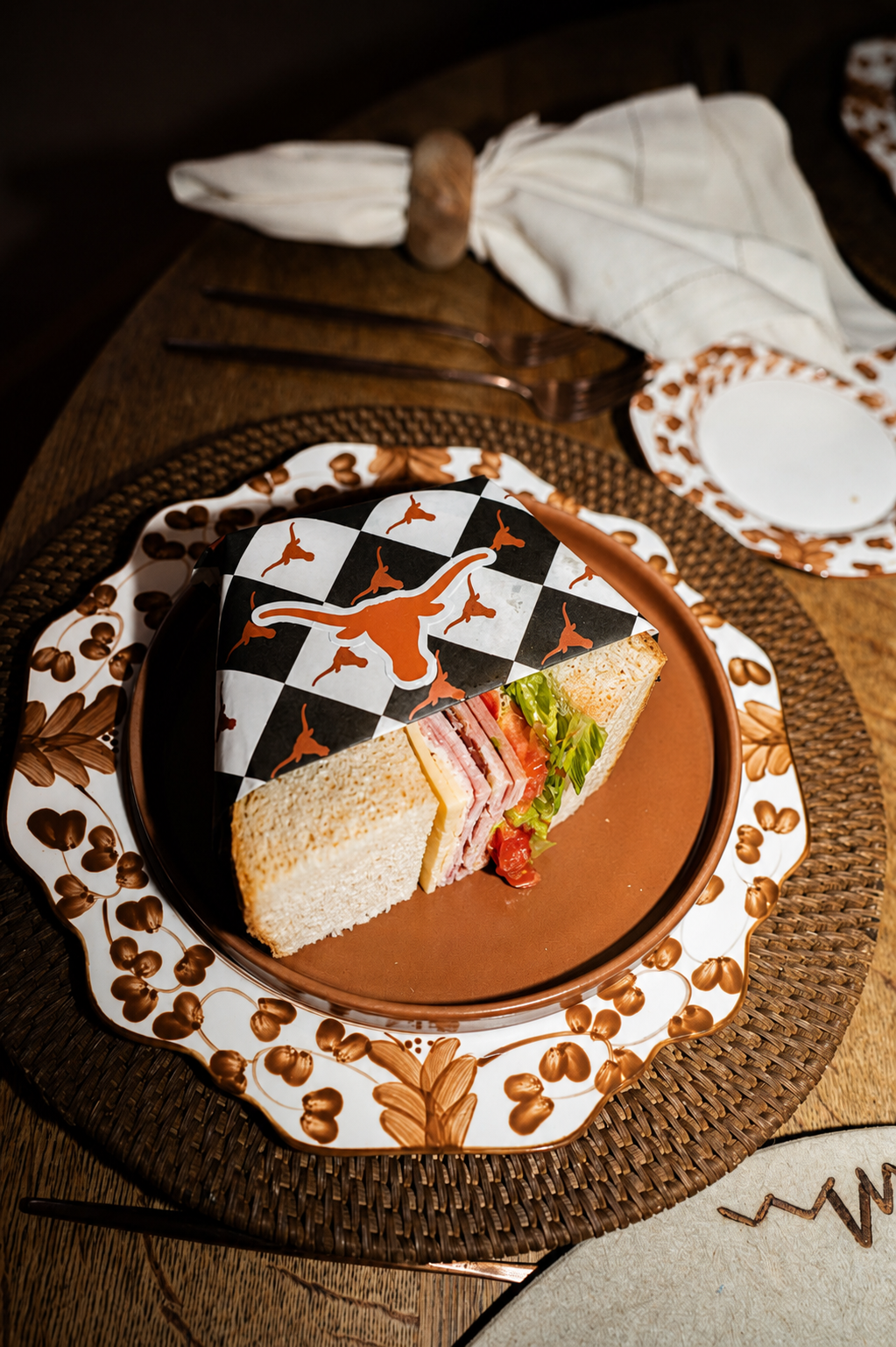 A platter with a Texas Longhorn logo and a checkered pattern, holding a sandwich with various meats, cheese, lettuce, and tomato, partially covered with themed paper.