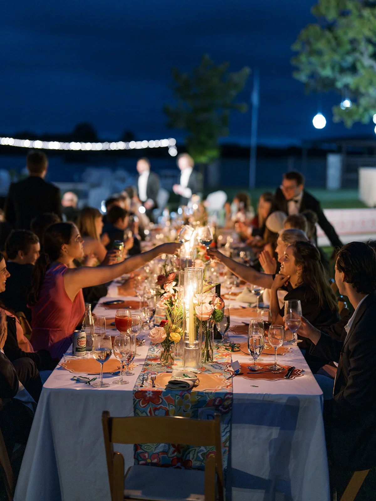 People celebrating at a long outdoor dinner table with floral decorations and candles at night.