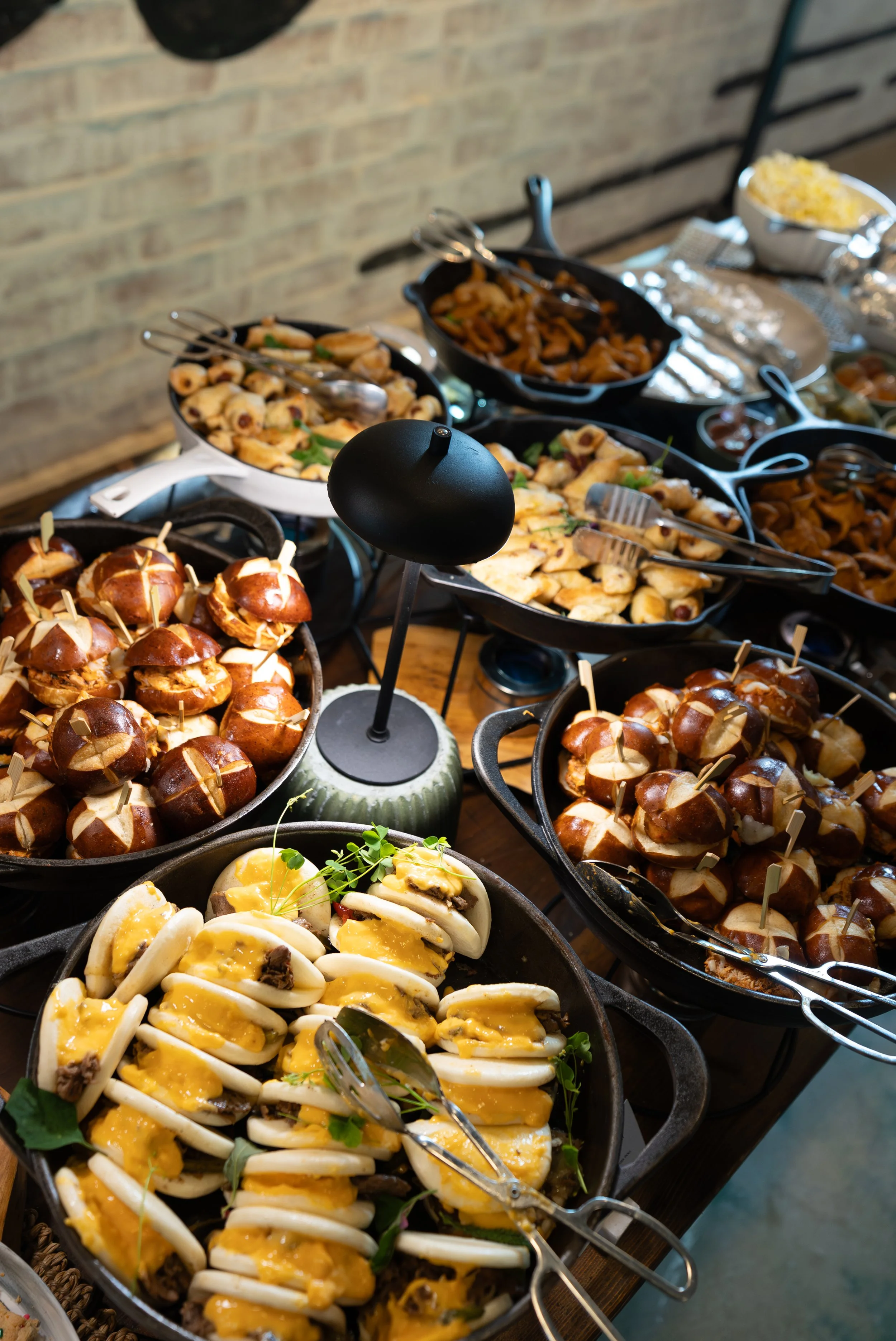 A buffet table with various dishes including stuffed pretzels, sliced boiled eggs with mustard and herbs, roasted vegetables, and other appetizers, arranged in cast iron skillets and bowls.