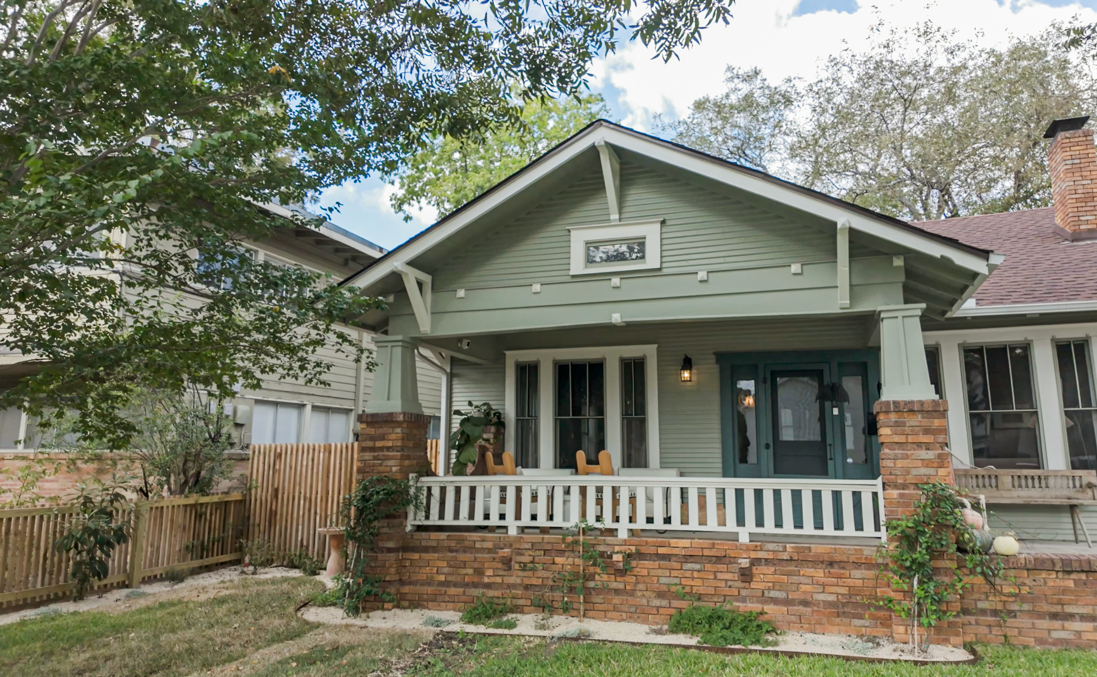 Front view of a light green house with white trim, a porch with white railing, and brick accents, surrounded by trees and a fence.