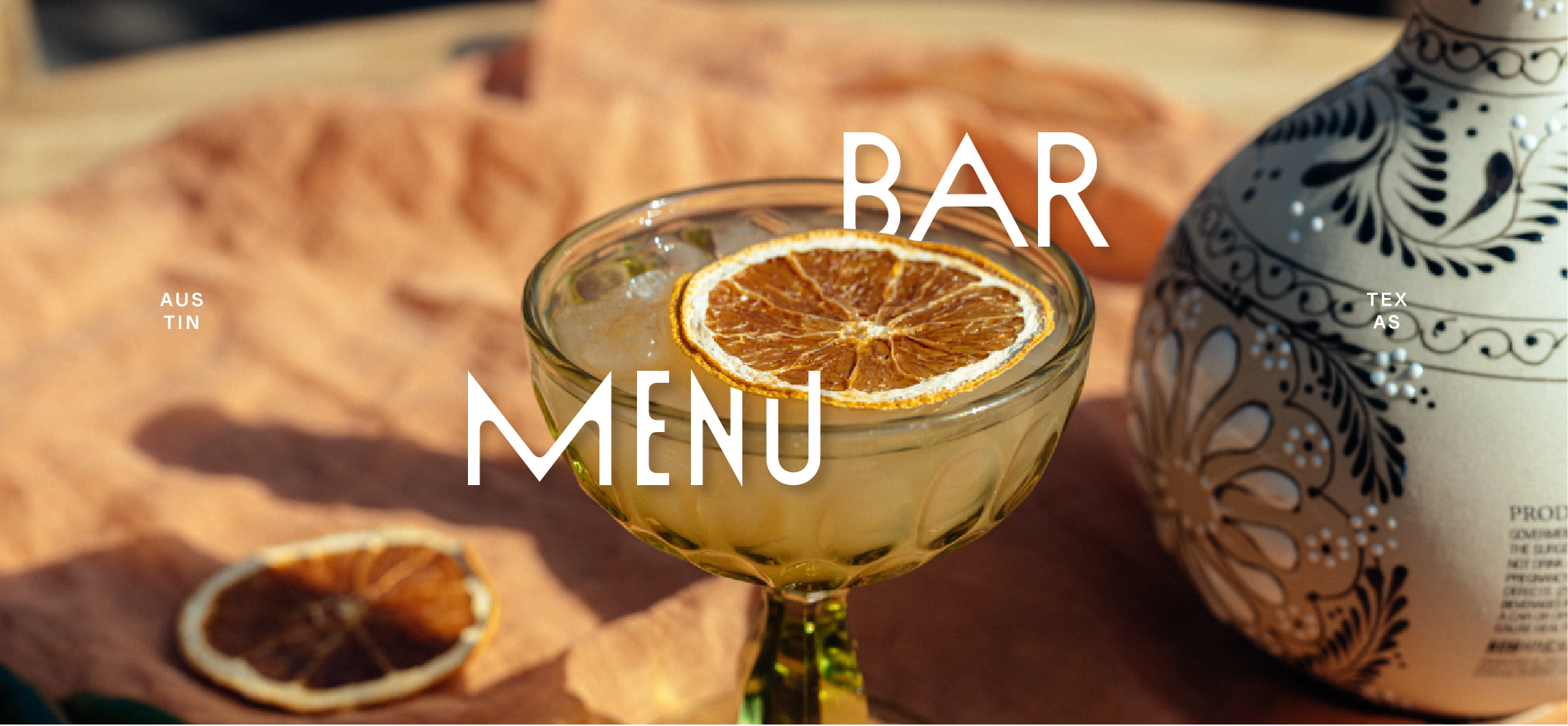 A cocktail glass with ice and a dried orange slice on top, set on a table with blurred background and decorative vase nearby.
