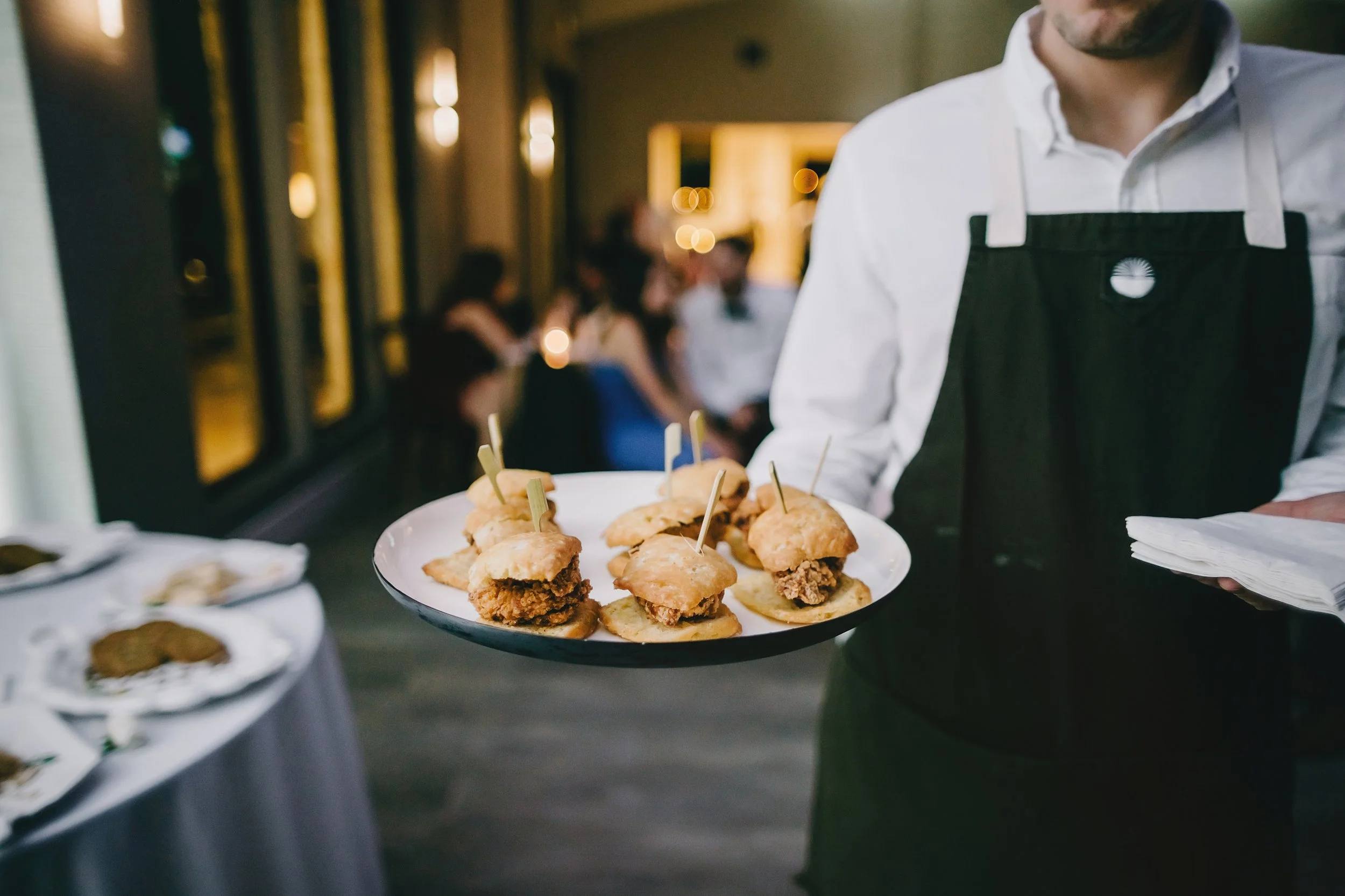 A waiter holding a plate of small sandwiches with fried chicken, with people dining in the background at a dimly lit restaurant.