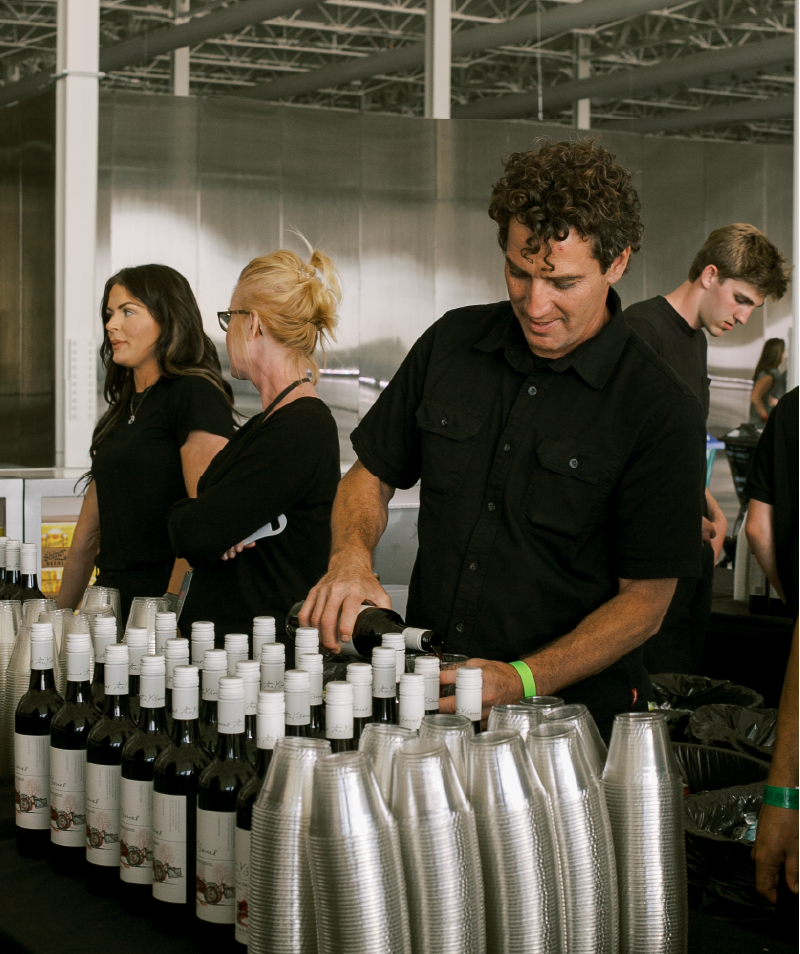 A man pouring wine into plastic cups at a wine tasting event, with several bottles of wine and stacked cups on the table.