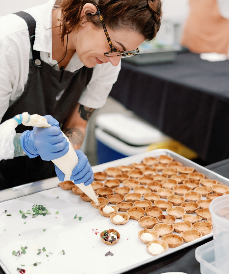 A woman in glasses and gloves is piping stuffing or frosting onto small pastry shells arranged on a tray.