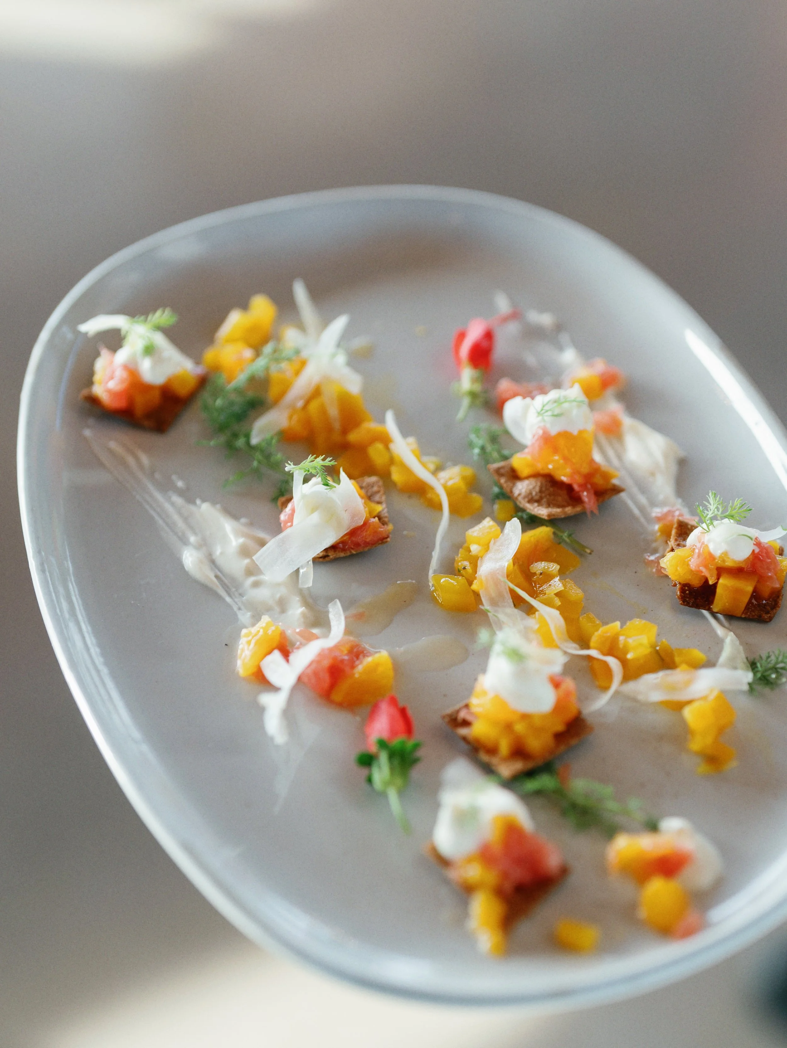 A close-up of a gray plate with small appetizers, including pieces of toast topped with diced vegetables, garnished with herbs, and drizzled with creamy sauce.