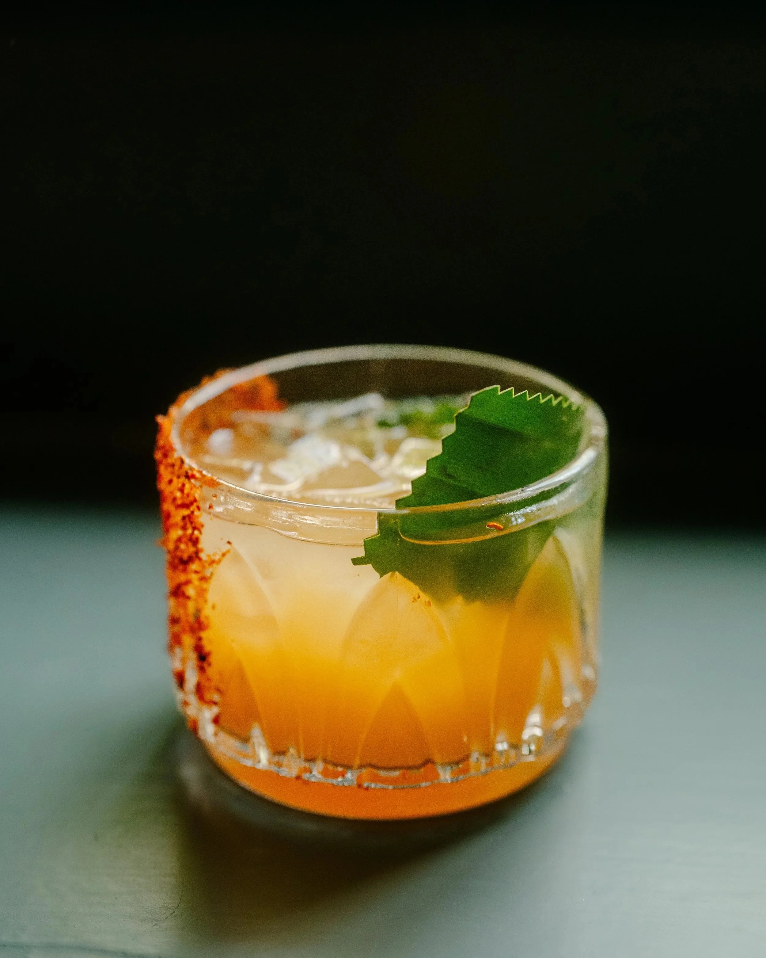 A cocktail drink in a transparent glass with a salted rim, garnished with a green leaf, ice cubes, lime slices, and decorative red and orange sugar or spice around the edge of the glass, set against a dark background.
