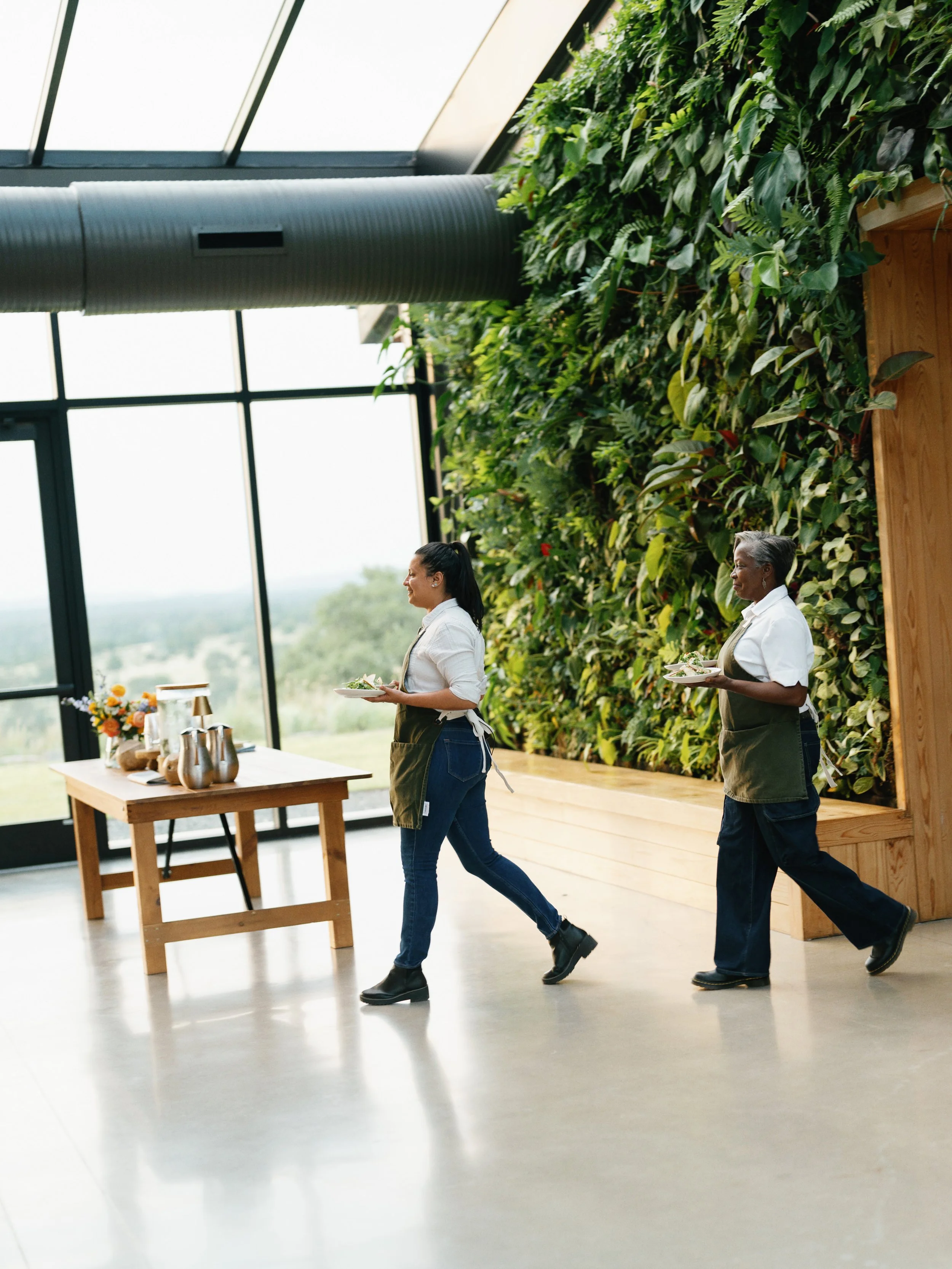 Two women walking in an indoor space with a green wall and large windows, holding plates of food.