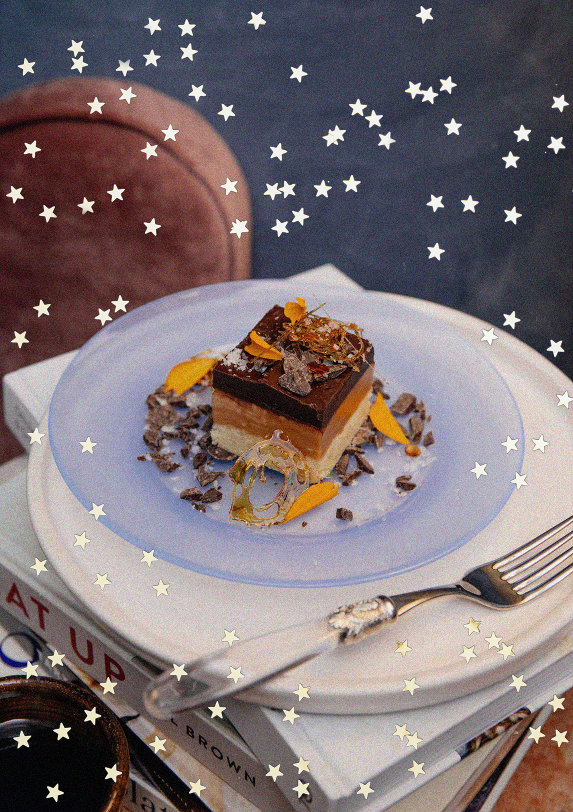 A layered chocolate and vanilla dessert on a white plate decorated with chocolate shavings, orange zest, and caramel, placed on top of a stack of books with a fork nearby, and a cup of coffee in the lower left corner.