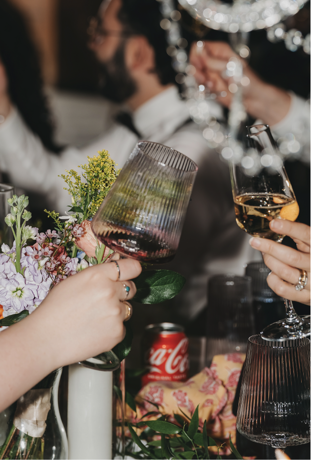 People clinking glasses with wine and champagne at a celebration with flowers, a Coke can, and a table setting.
