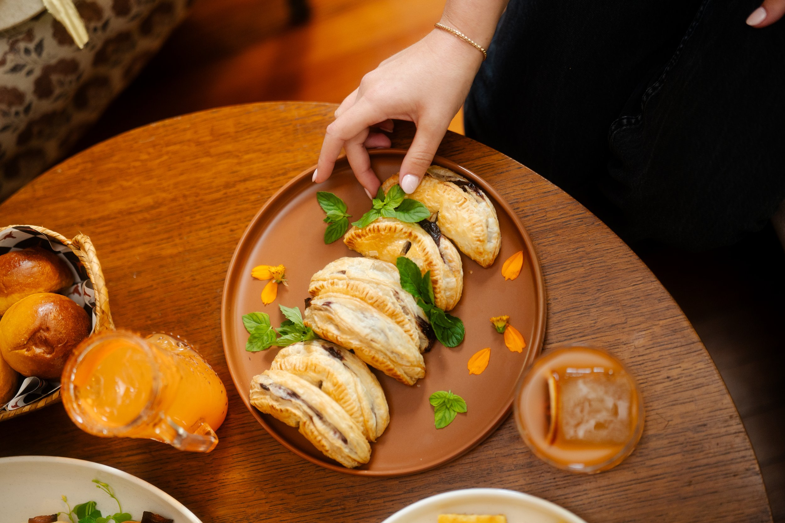 A plate of puff pastry turnovers garnished with fresh basil leaves and orange flower petals on a wooden table, with glasses of orange juice nearby.