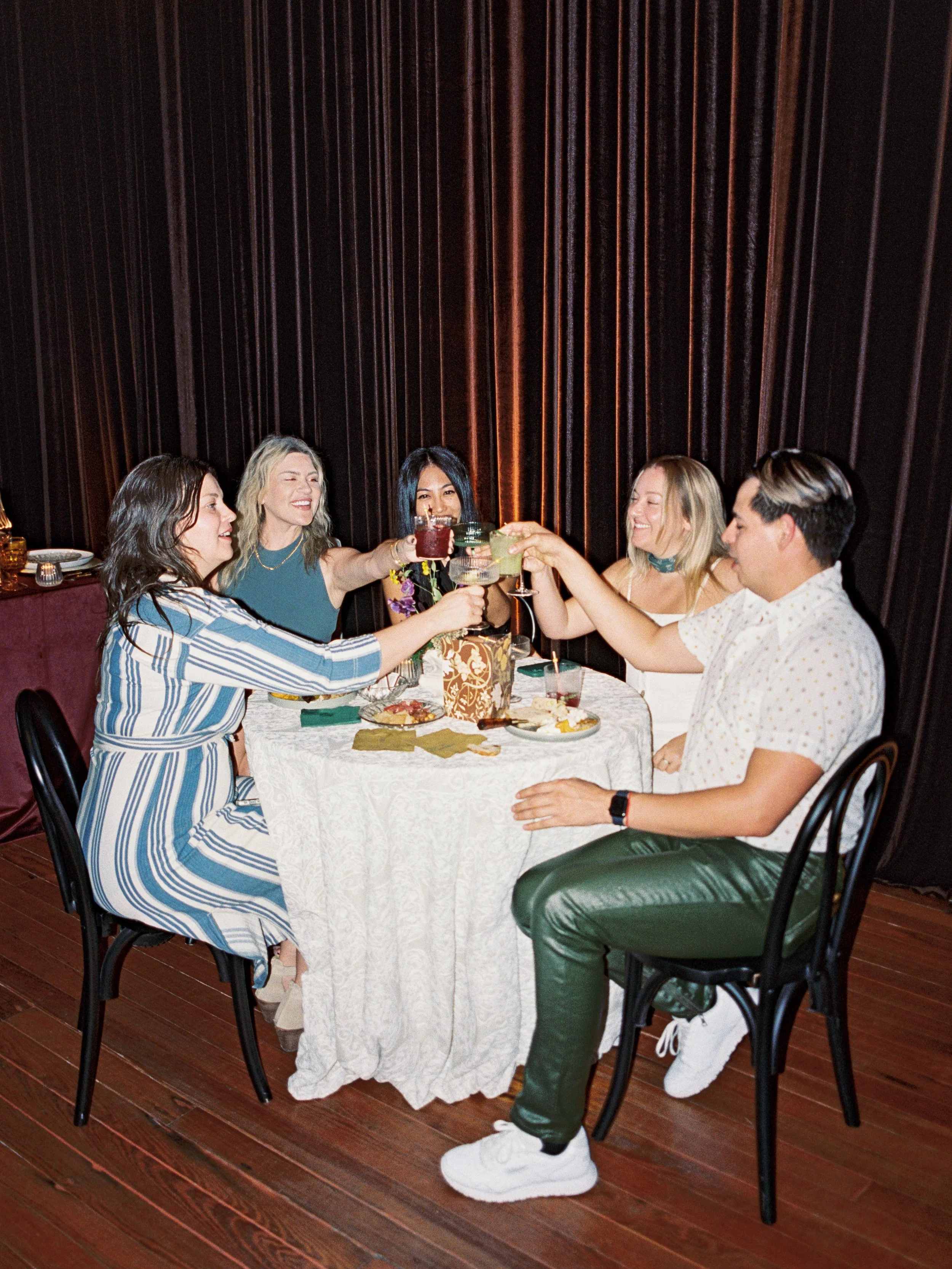 Group of six friends sitting around a table, toasting drinks, celebrating, with food and drinks on the table, against a dark curtain backdrop.