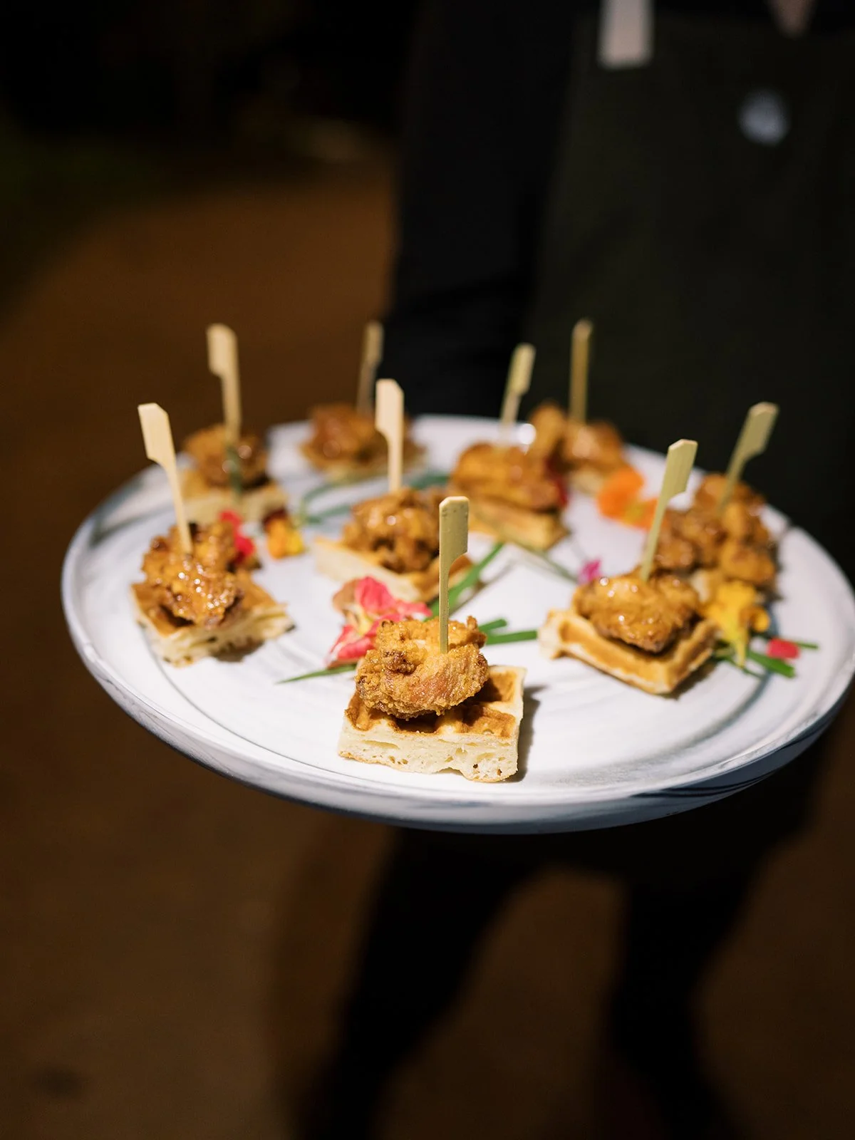 A tray of bite-sized appetizers with small pieces of fried chicken on bread, each with a toothpick, served on a white round platter decorated with flowers and greenery.