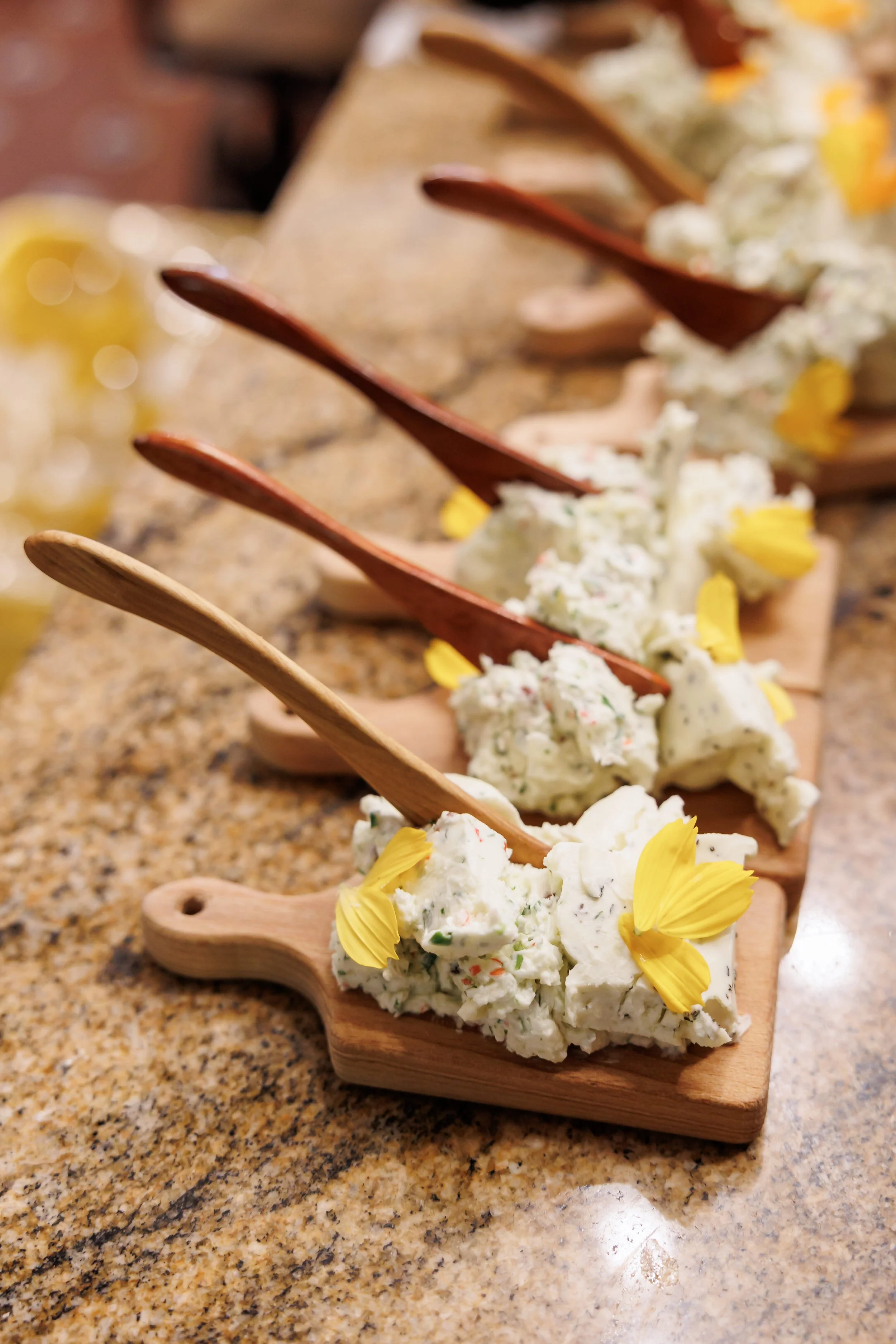 A row of cheese spreads topped with yellow flower petals, served on small wooden boards with cheese knives on a granite countertop.