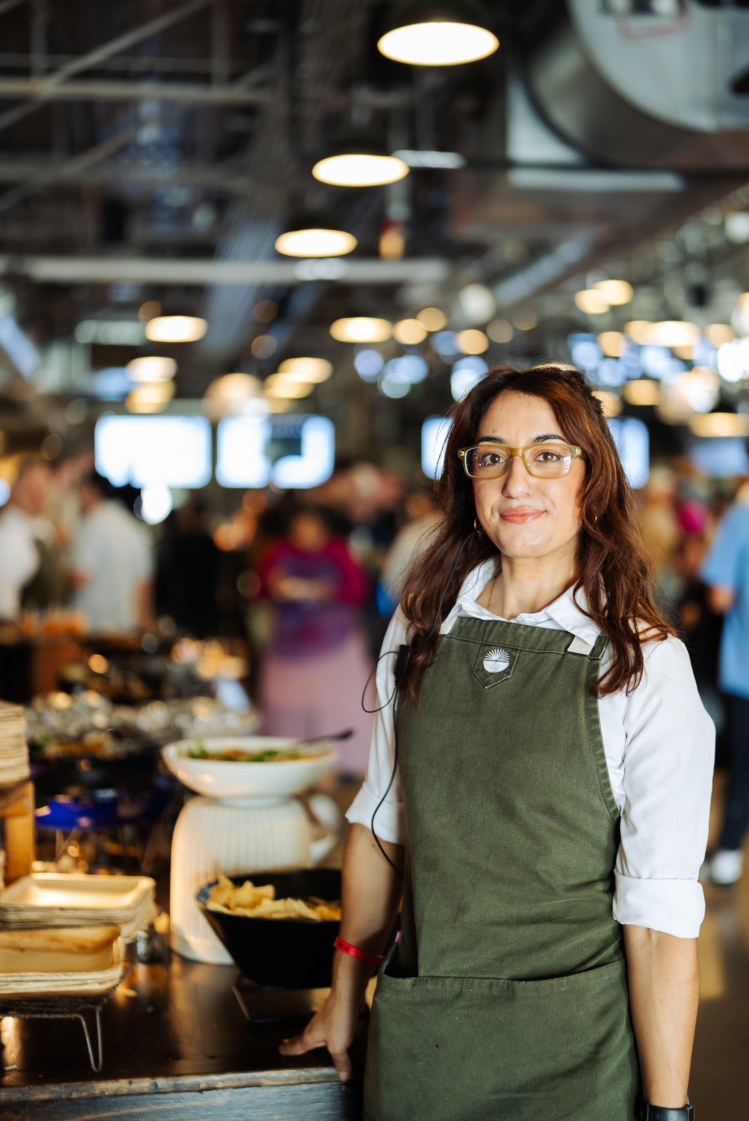 A woman with long dark hair and glasses wearing a white shirt and green apron, standing in a busy food market or restaurant, looking at the camera.