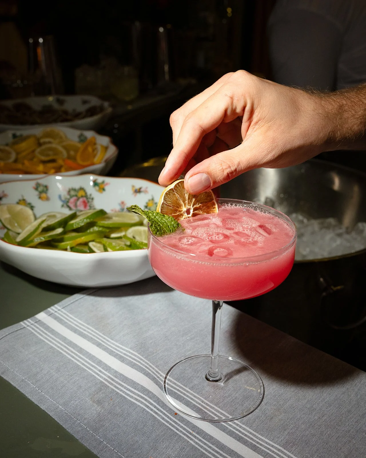 Hand garnishing a pink cocktail with a lemon slice and mint leaf in a coupe glass. There are bowls of lemon and lime wedges in the background.