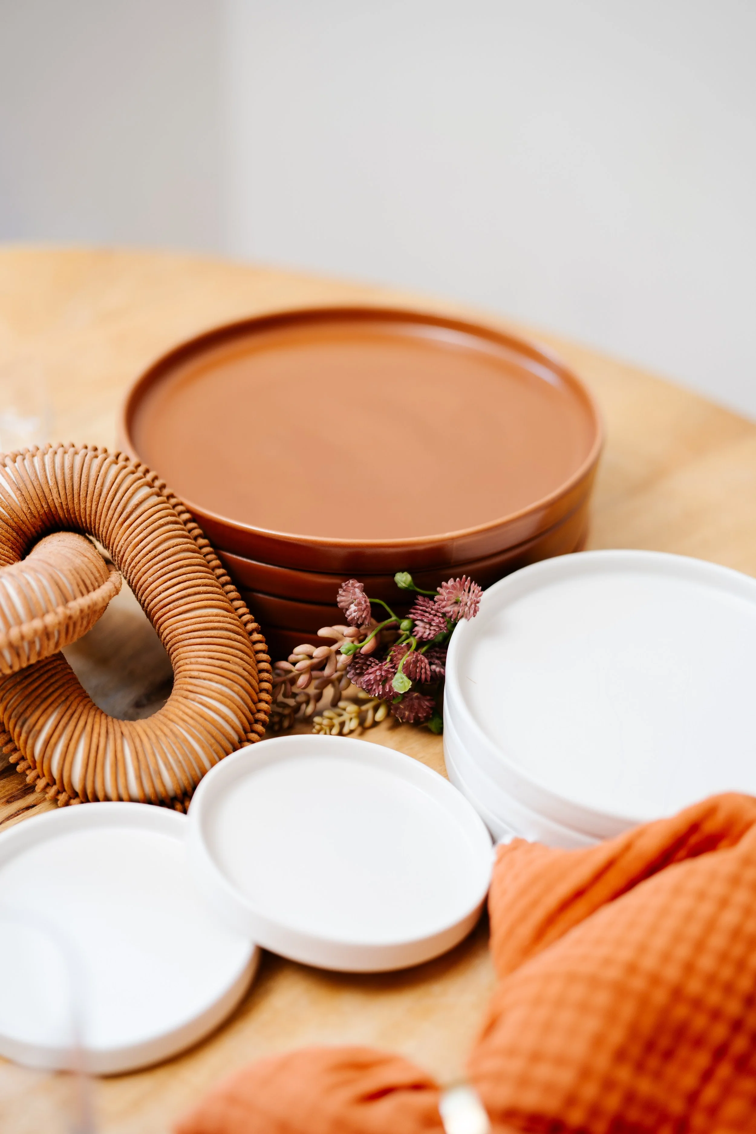 Arrangement of empty white bowls, clay pots, a wicker basket, and a small bundle of pink and purple flowers on a wooden surface.