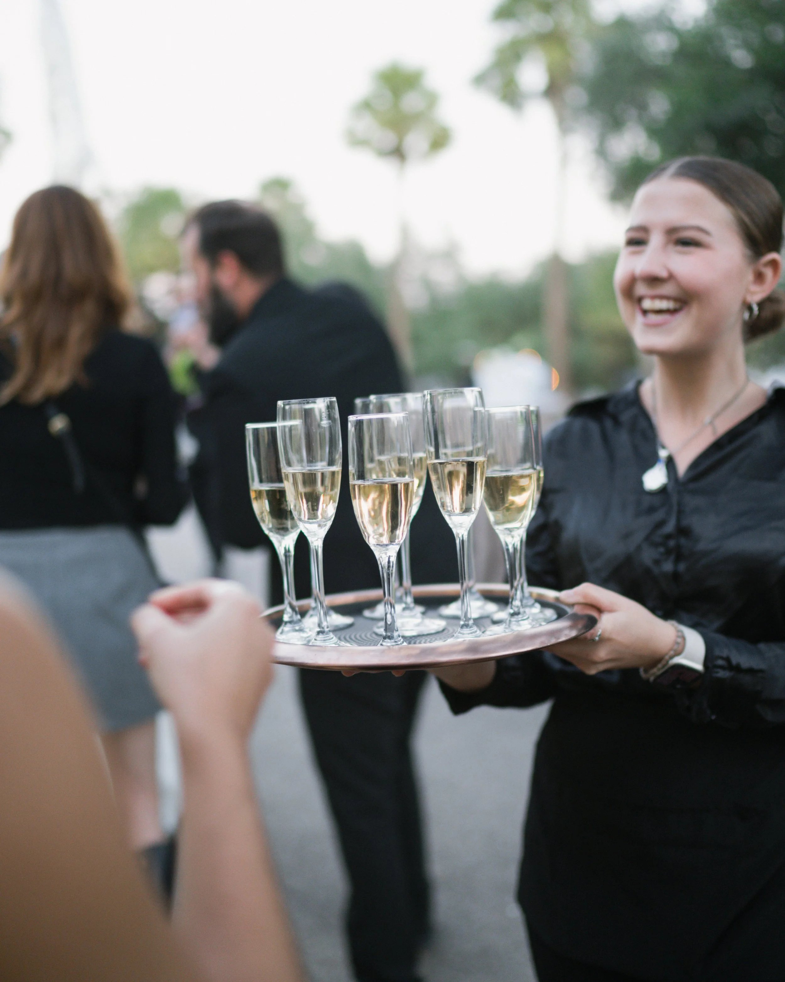 A woman in black holding a tray with several glasses of champagne or sparkling wine. In the background, two people are blurred, talking to each other at what appears to be an outdoor event with trees and balloons.