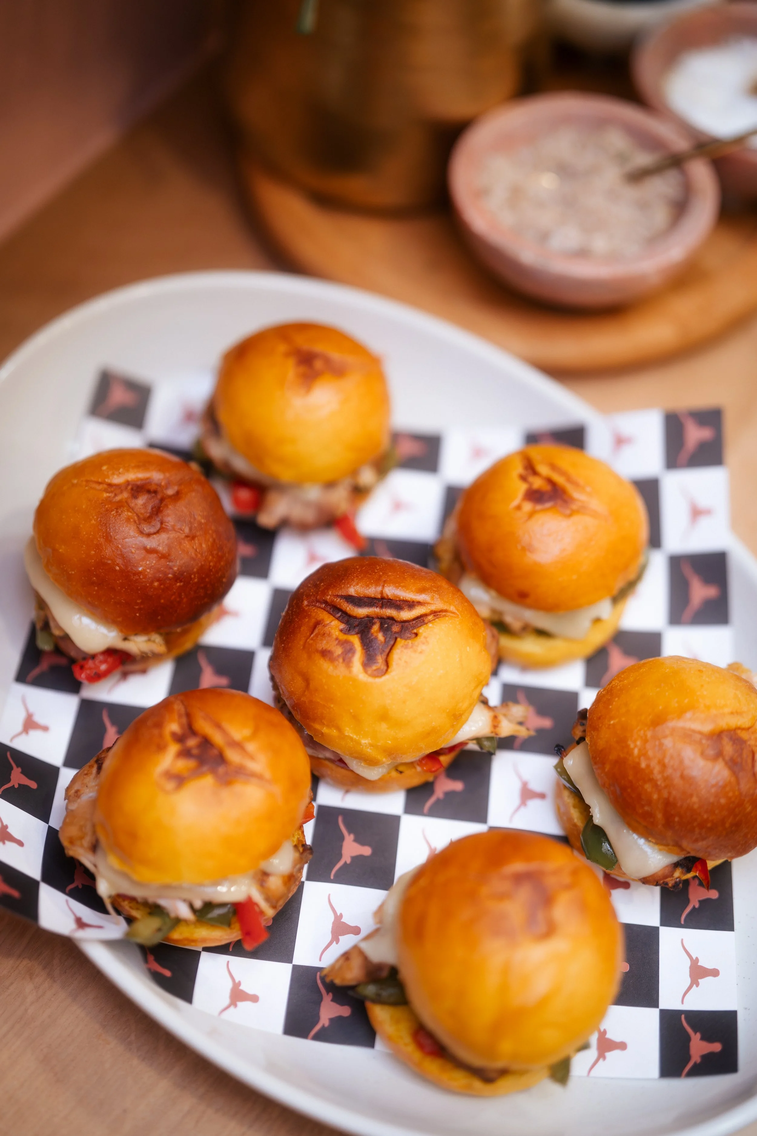 Mini slider sandwiches with buns topped with grilled markings, filled with cheese, meat, and vegetables, arranged on a black and white checkered tray with a pink logo pattern. Background shows bowls with sauces or condiments.