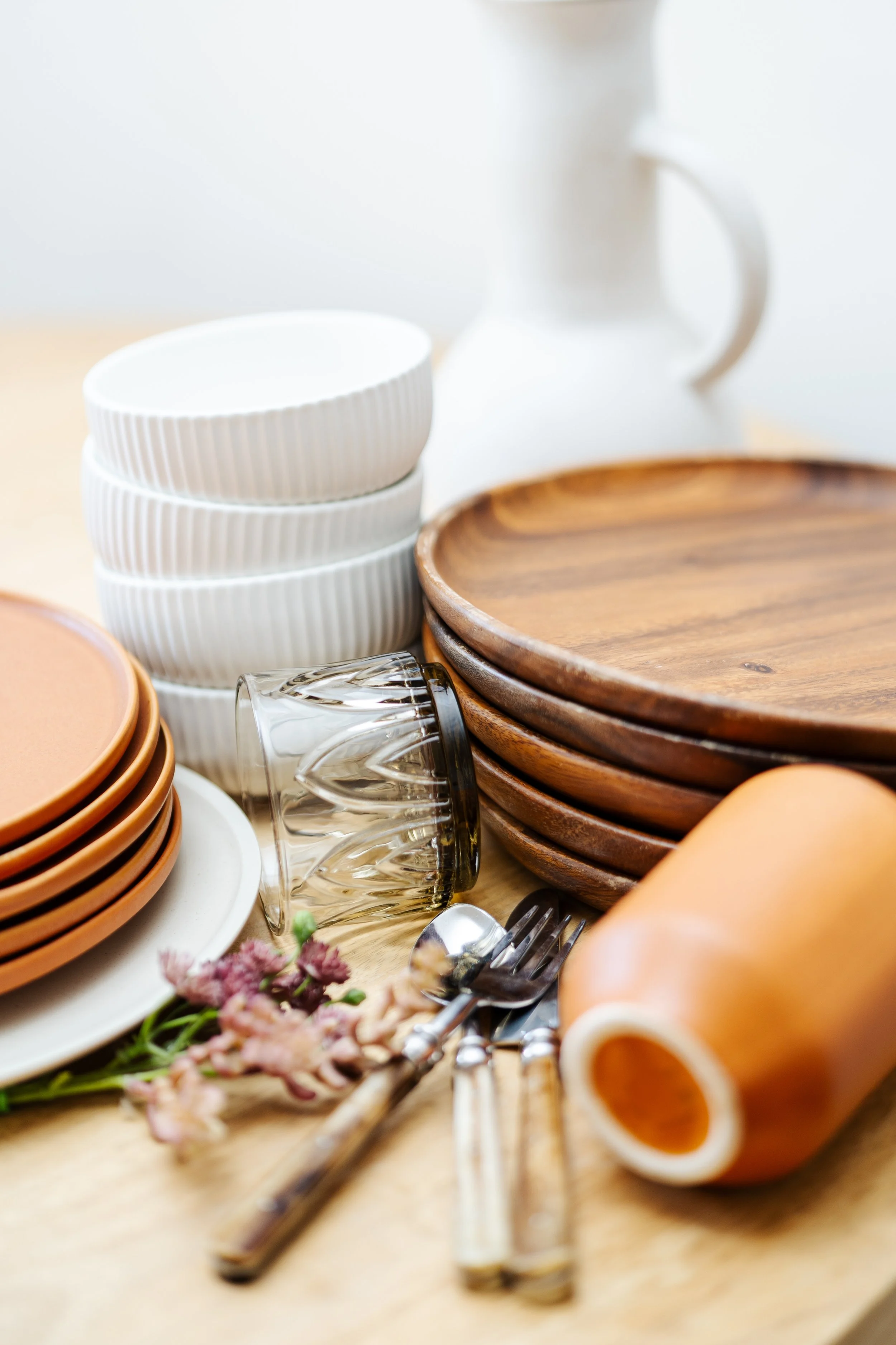 Stacked white bowls, brown plates, clear glass goblet, and orange ceramic cups with a floral decoration in the foreground on a light wooden table.