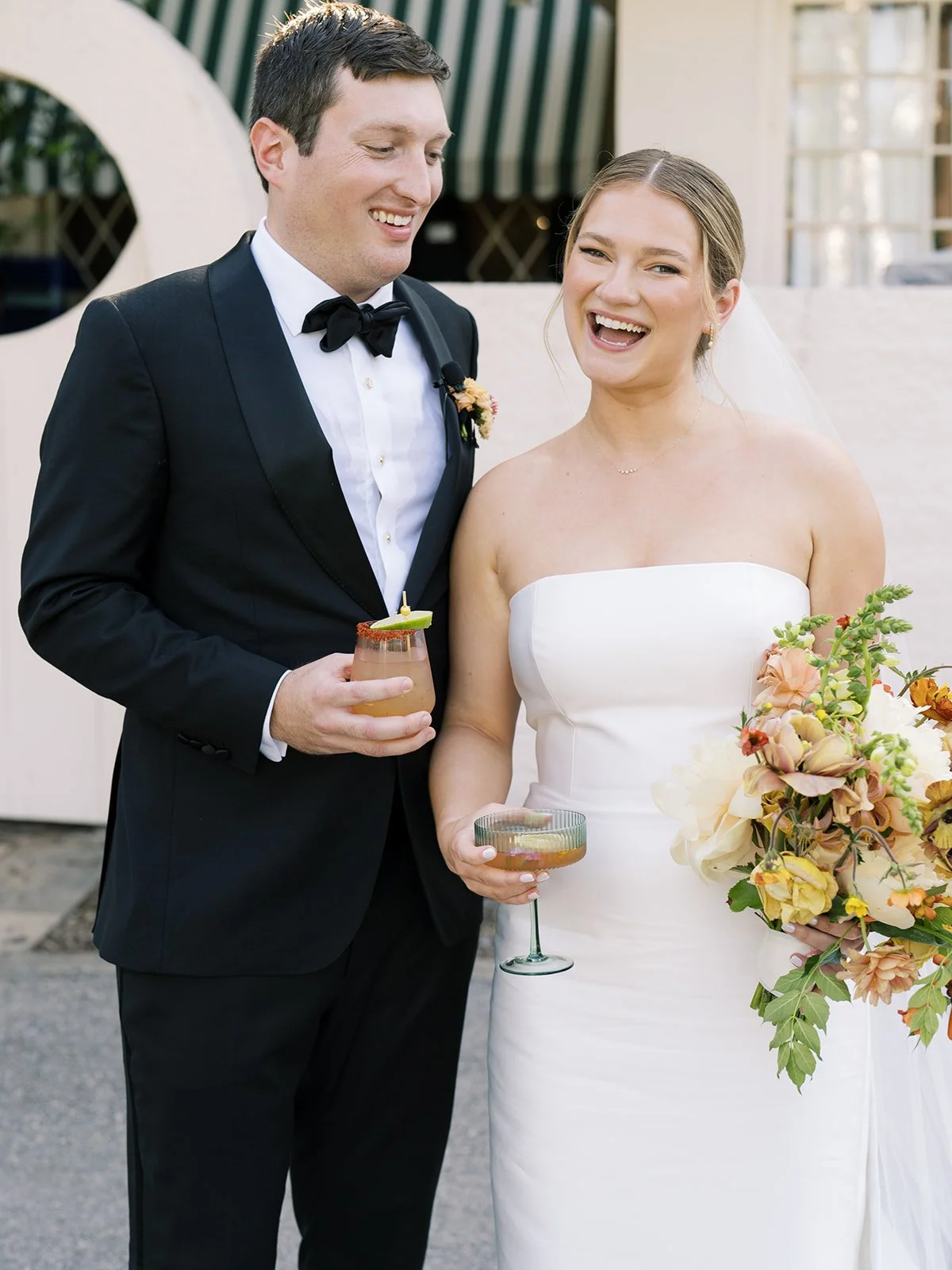A newlywed couple smiling and holding drinks during their wedding celebration. The groom is wearing a black tuxedo with a bow tie, and the bride is in a strapless white wedding dress holding a bouquet of flowers.