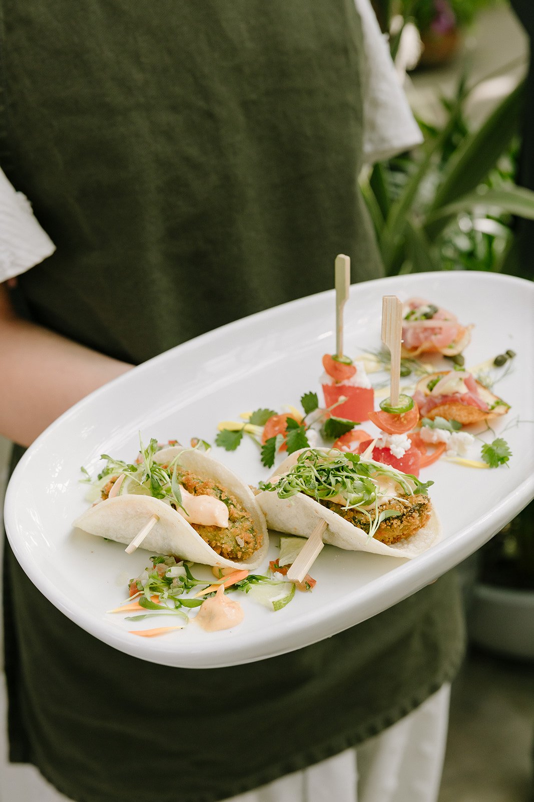 A person holding a white oval platter with three taco-style appetizers topped with microgreens, along with small skewers of tomato and cheese, garnished with herbs.