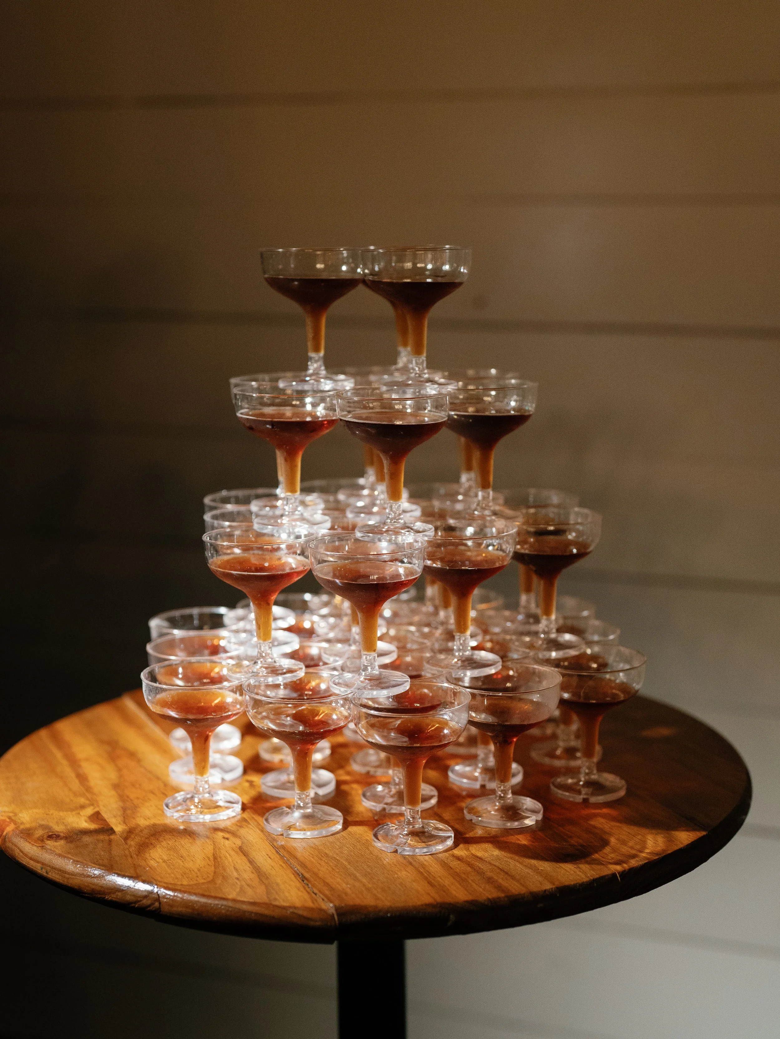 A pyramid of stacked cocktail glasses filled with dark brown and reddish beverages on a wooden table.