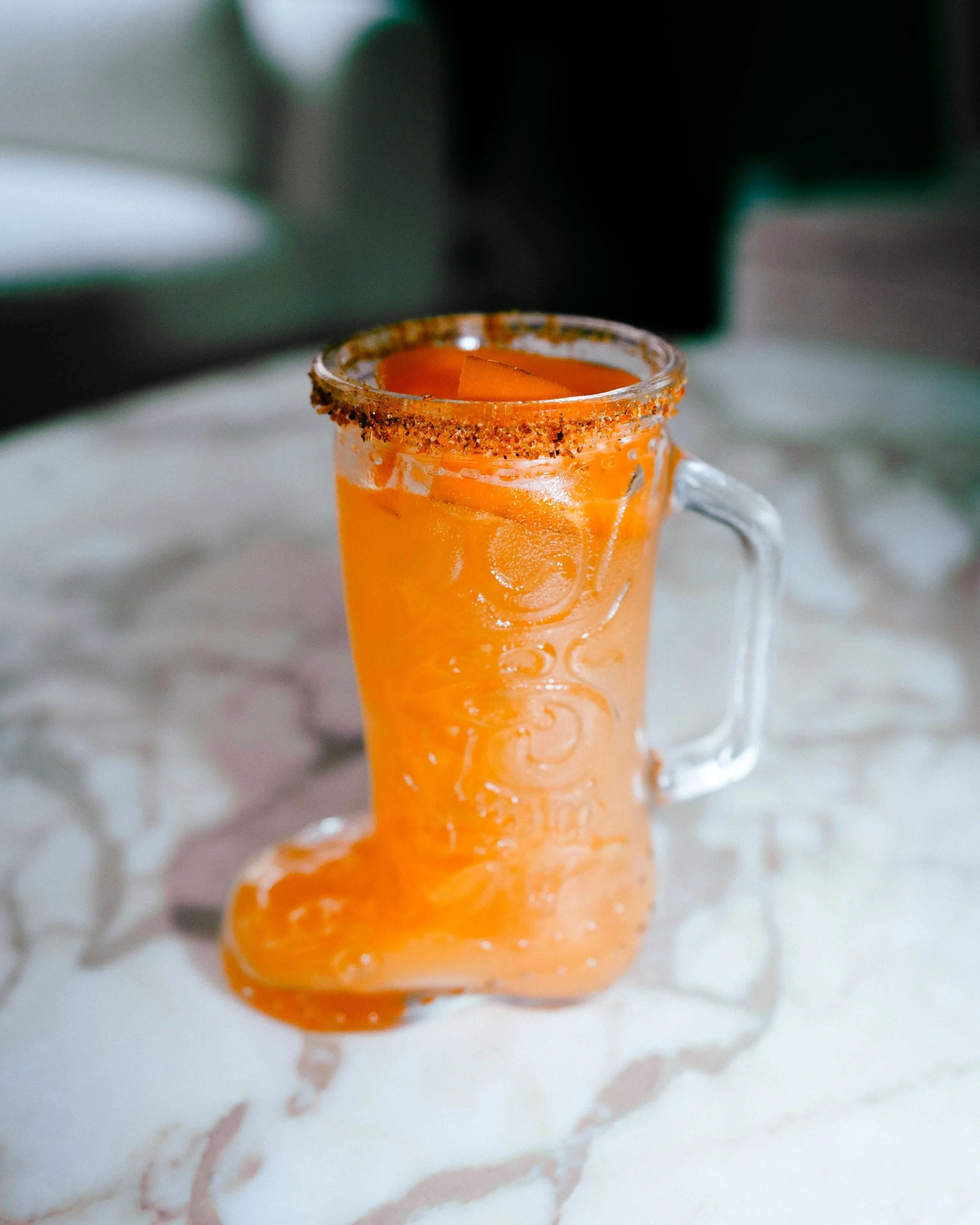 A glass mug with an orange-colored beverage, garnished with a slice of orange, and the rim coated with chili powder or spice.