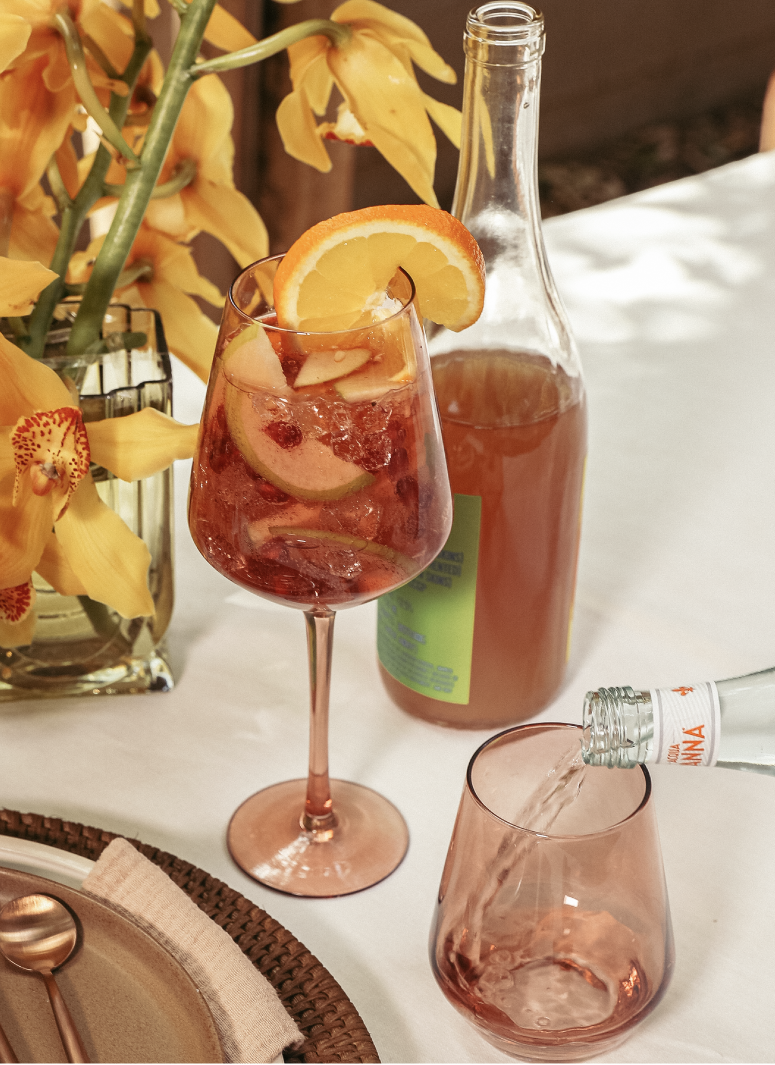 A glass of pink drink with ice and lemon slices, a bottle of pink beverage, and a glass pouring a similar drink, on a white table with a flower arrangement in the background.