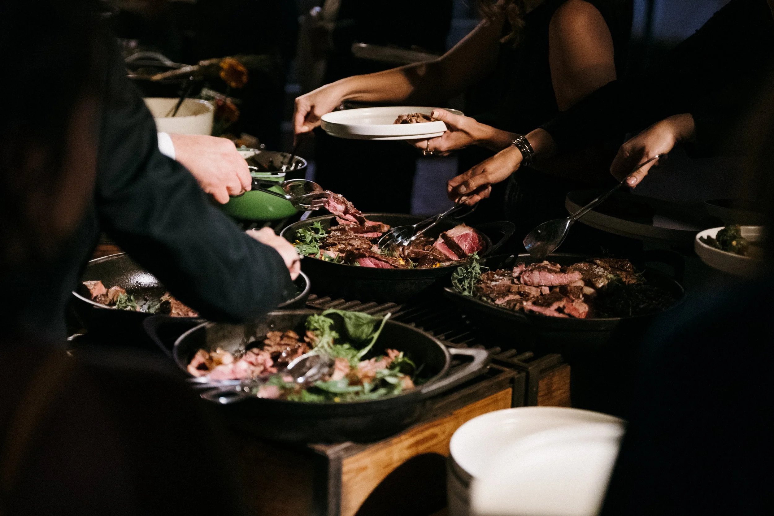 People serving themselves food from a buffet with various meats and greens at a dining event.