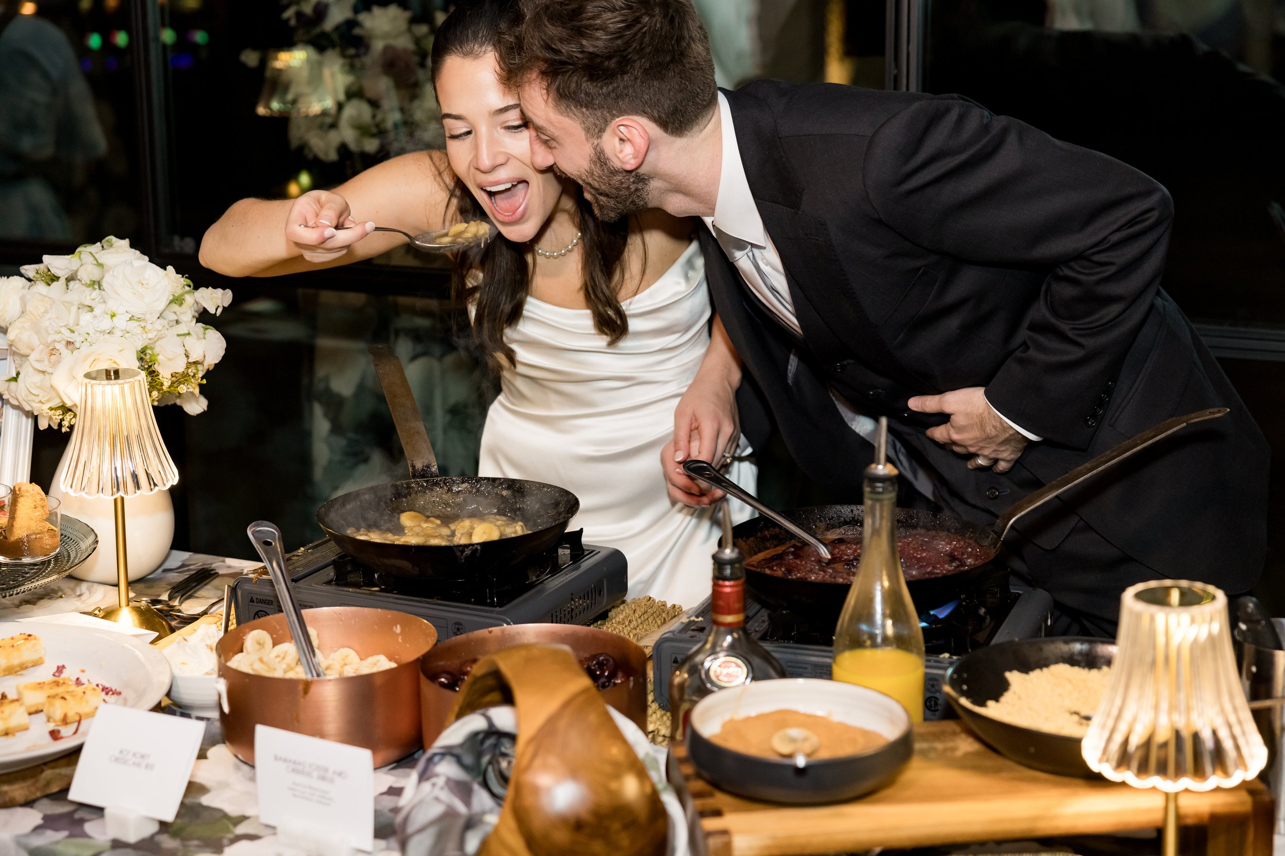 A woman in a white dress and a man in a black suit are cooking together at a table with various dishes, sauces, and a lamp, sharing a moment of joy and laughter.