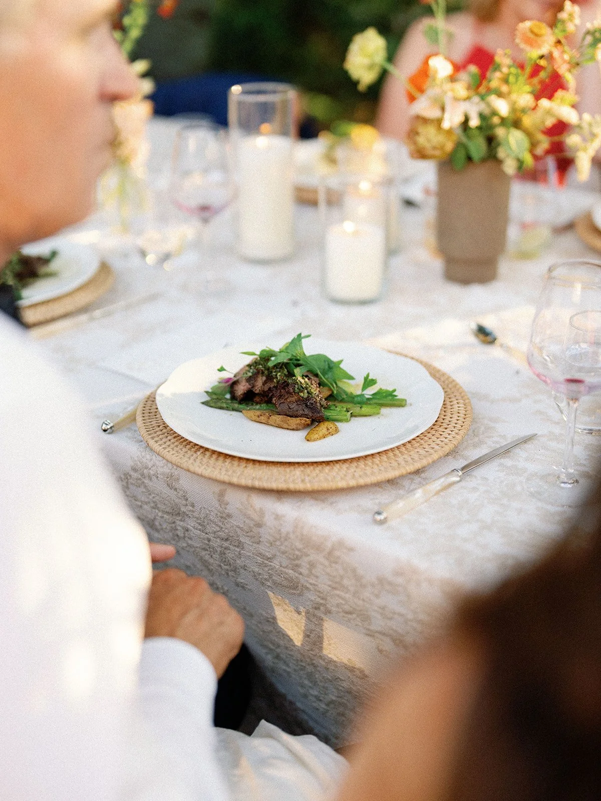 A plated meal with meat and greens on a dining table during a gathering, decorated with candles and flowers.