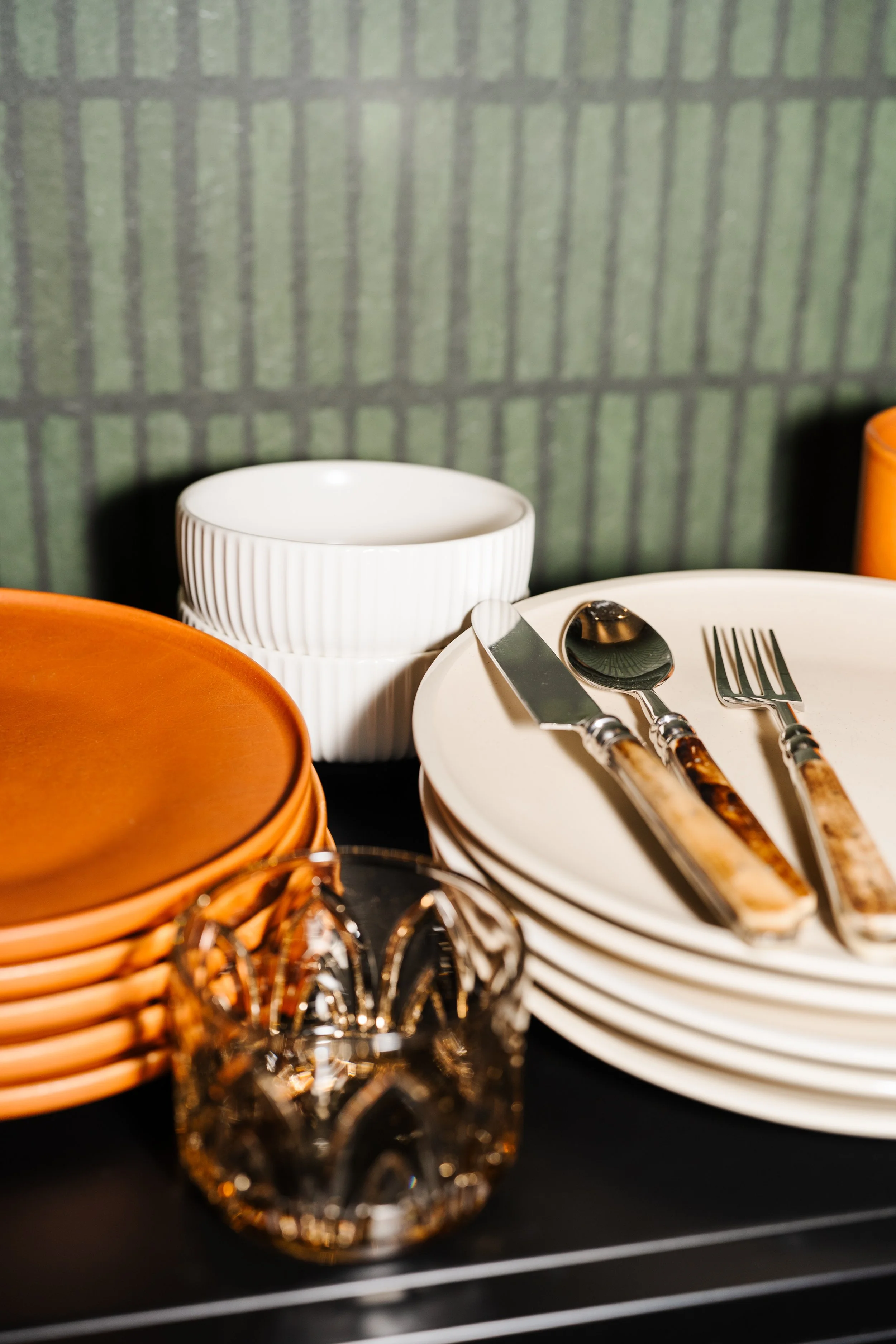 A close-up view of stacked plates, a white bowls, and cutlery on a black surface with a green patterned background.