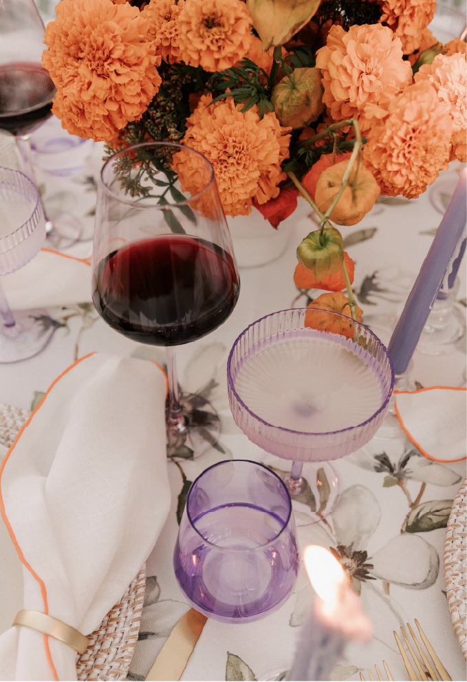 A table setting featuring a floral centerpiece with orange carnations and other flowers, a glass of red wine, and various purple-tinted glassware, including a wine glass, a cocktail glass, and a tumbler, along with a white napkin with orange trim, gold-colored utensils, and a lit candle.
