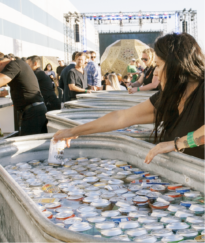 People at an outdoor event with a stage in the background are collecting and recycling aluminum cans in large silver bins.
