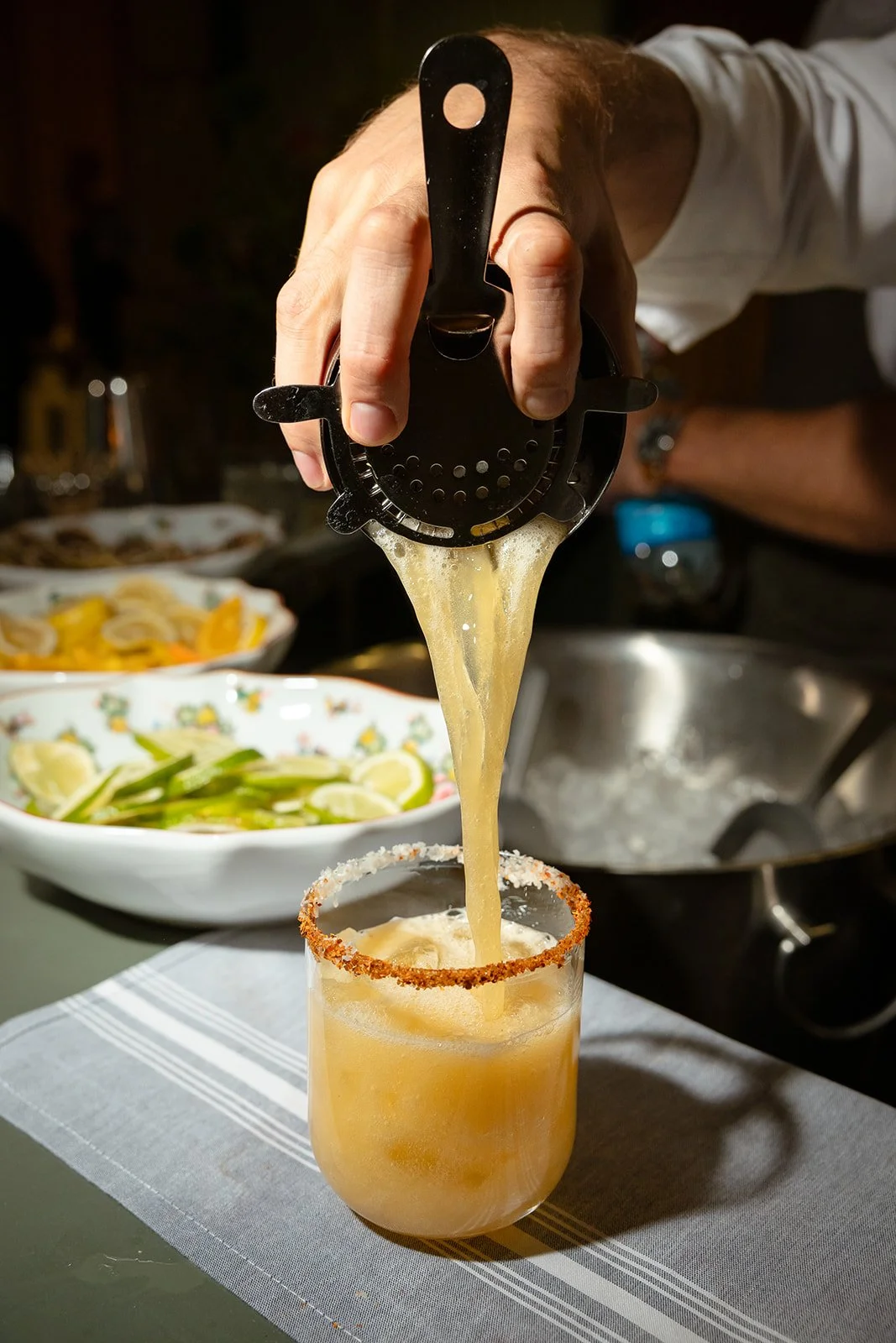 A person using a presser to pour a yellow beverage into a glass with a salted rim, with bowls of lemon slices and other ingredients in the background.