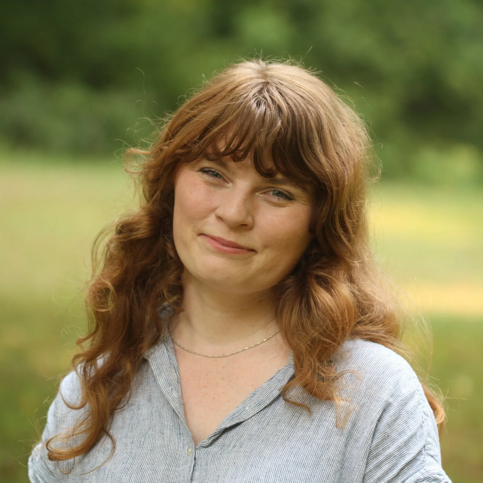 A person with white skin, long red hair and bangs looks at the camera and smiles.