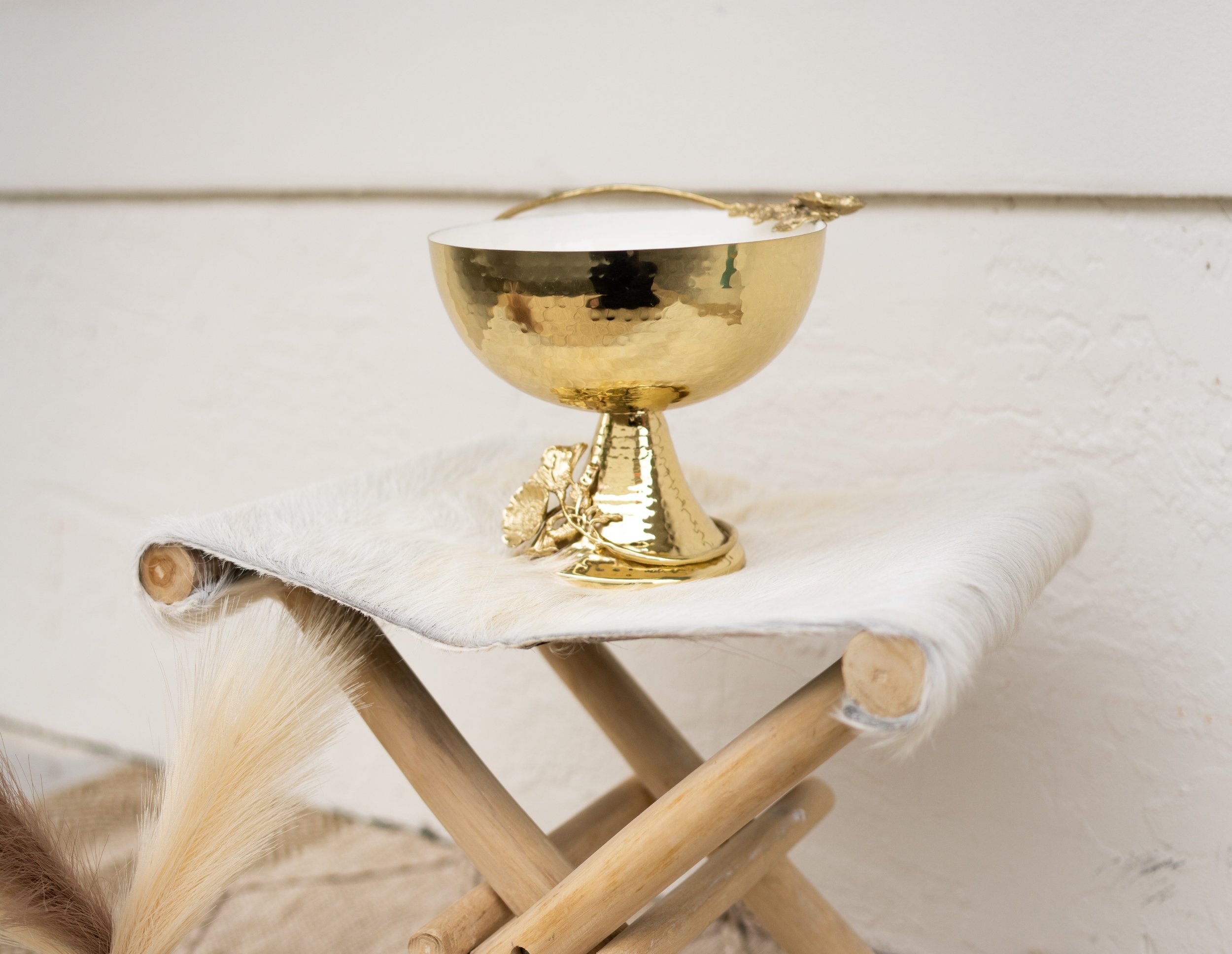 The Noora Golden Flora Bowl rests on a white fur-textured stool, showcasing its gleaming gold finish and sophisticated silhouette, exemplifying luxury in home decor.
