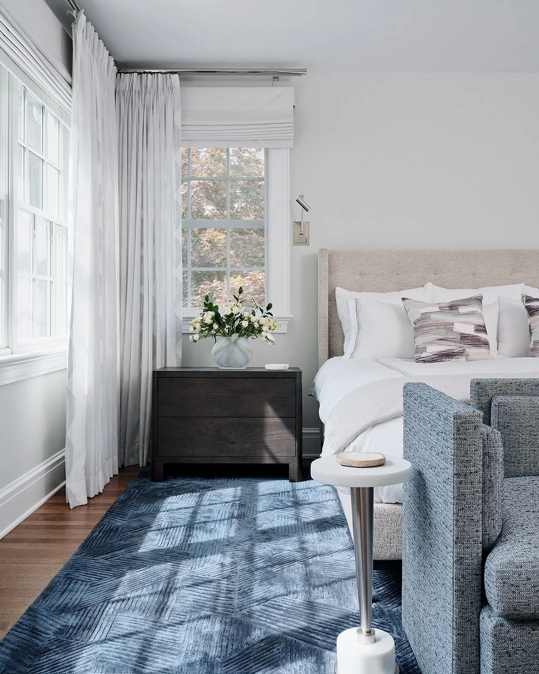 Swipe for the thoughtful details that bring the Scarsdale Colonial primary bedroom together: 

a neutral upholstered bed, polished nickel sconces, a custom indigo rug, layered window treatments, a textured bench, dark-stained furnishings, and an octa