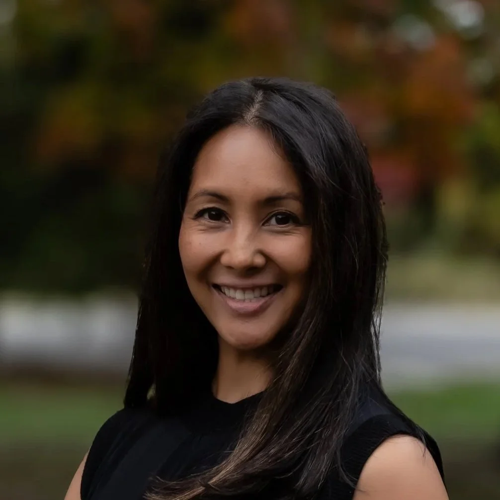 A woman with shoulder-length black hair smiling outdoors near a tree with purple flowers, green grass, and other trees in the background.